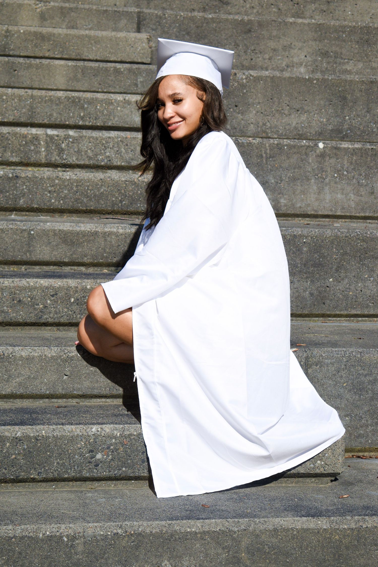 Graduate in white cap and gown sits on concrete steps during outdoor photo session.