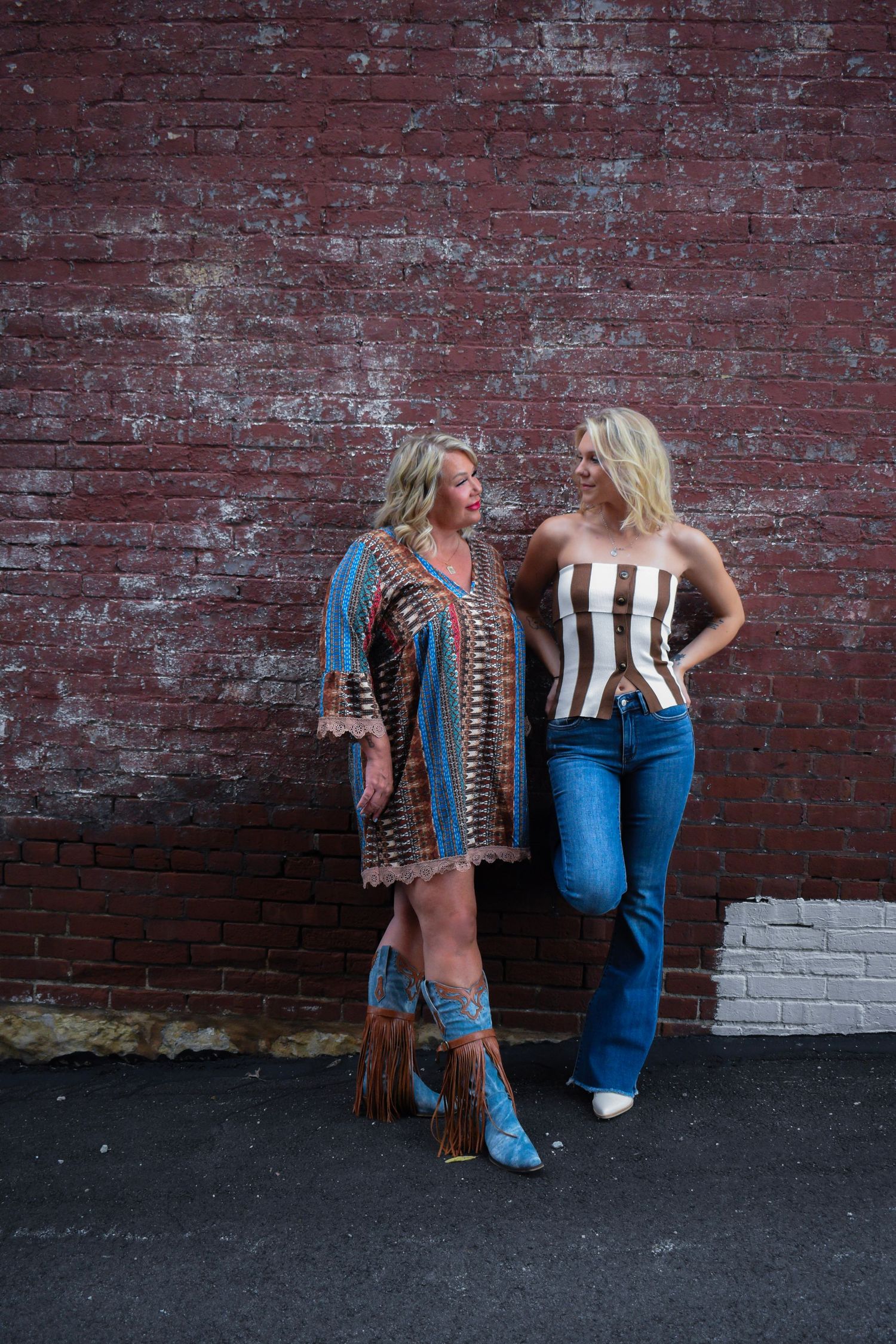 Two people posing against a rustic brick wall wearing contrasting casual and bohemian outfits.