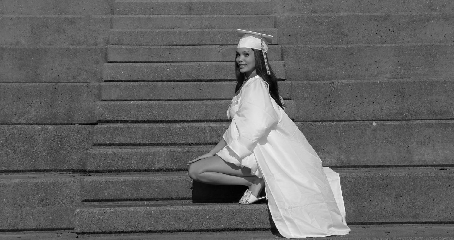 Graduate in white regalia sitting on concrete steps in black and white photography.