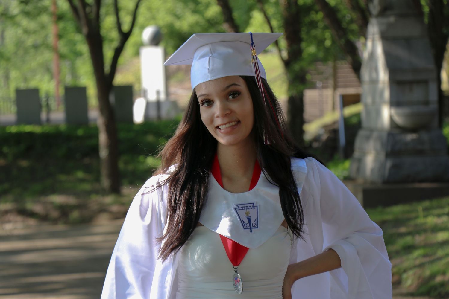 Graduate in white academic regalia smiling on tree-lined campus path.
