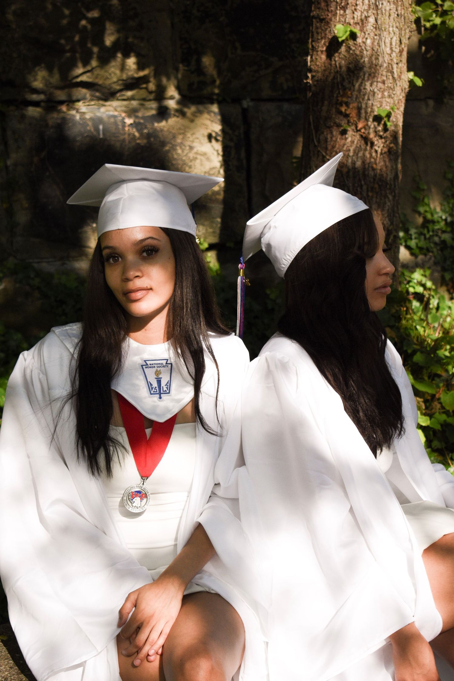 Two graduates in white regalia sitting back-to-back in a wooded setting.