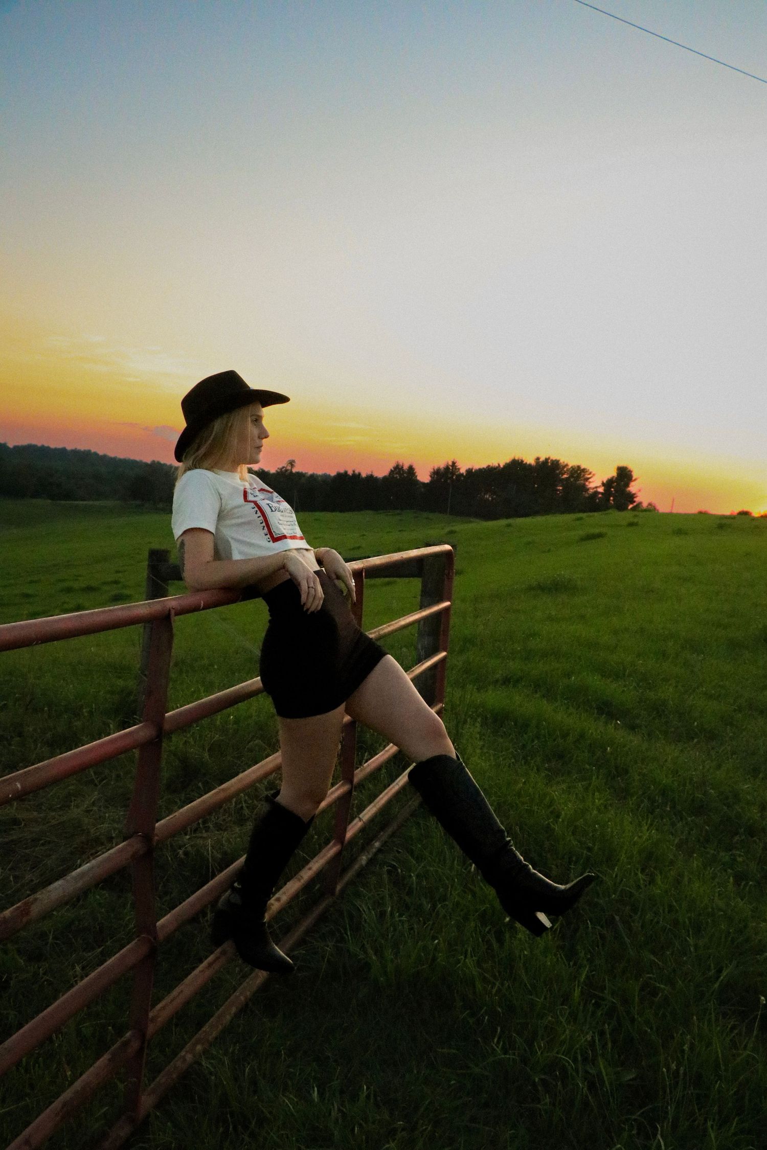 Silhouette against sunset leaning on wooden fence in rural field setting.