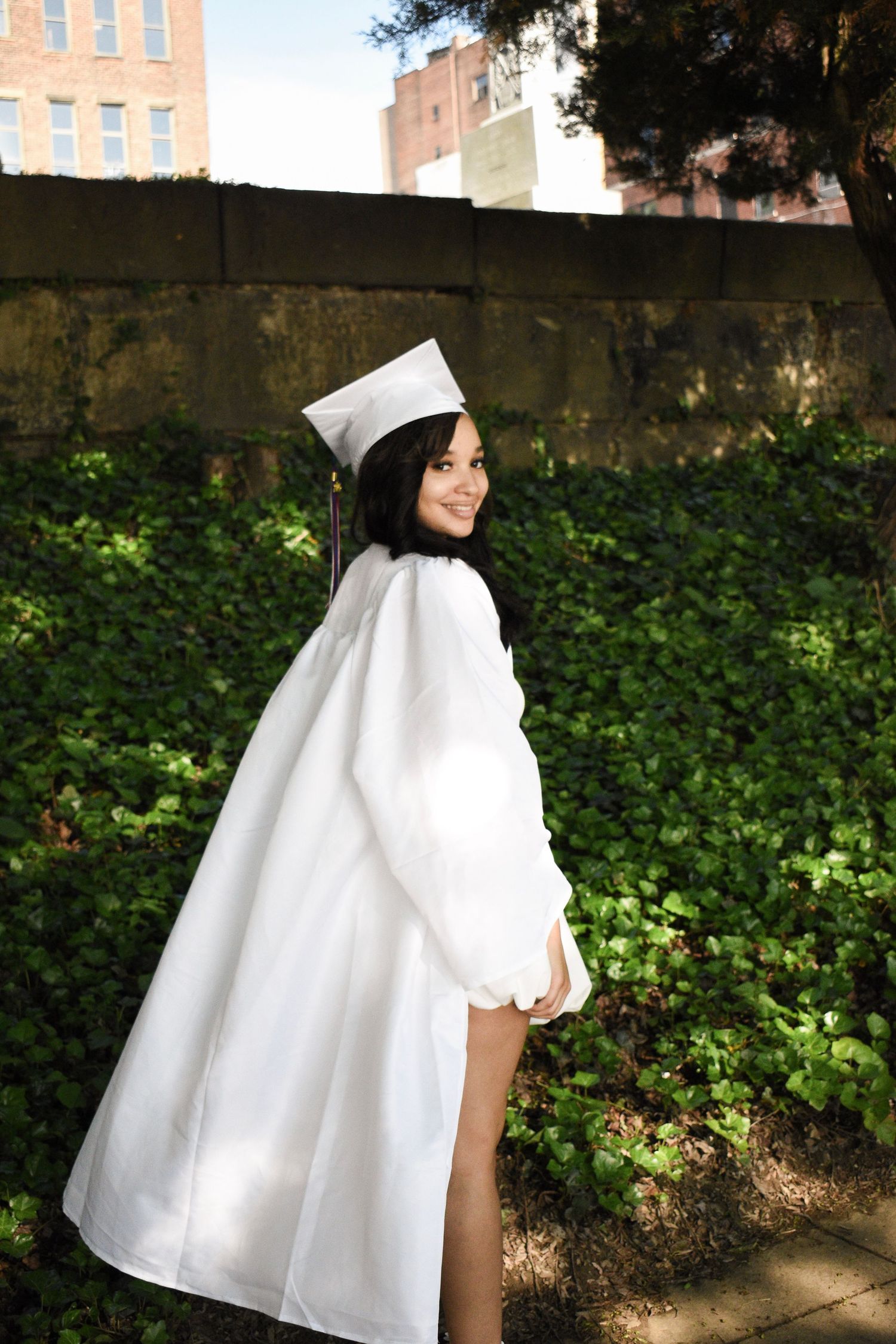Graduate in white cap and gown smiles over shoulder in sunny garden setting.