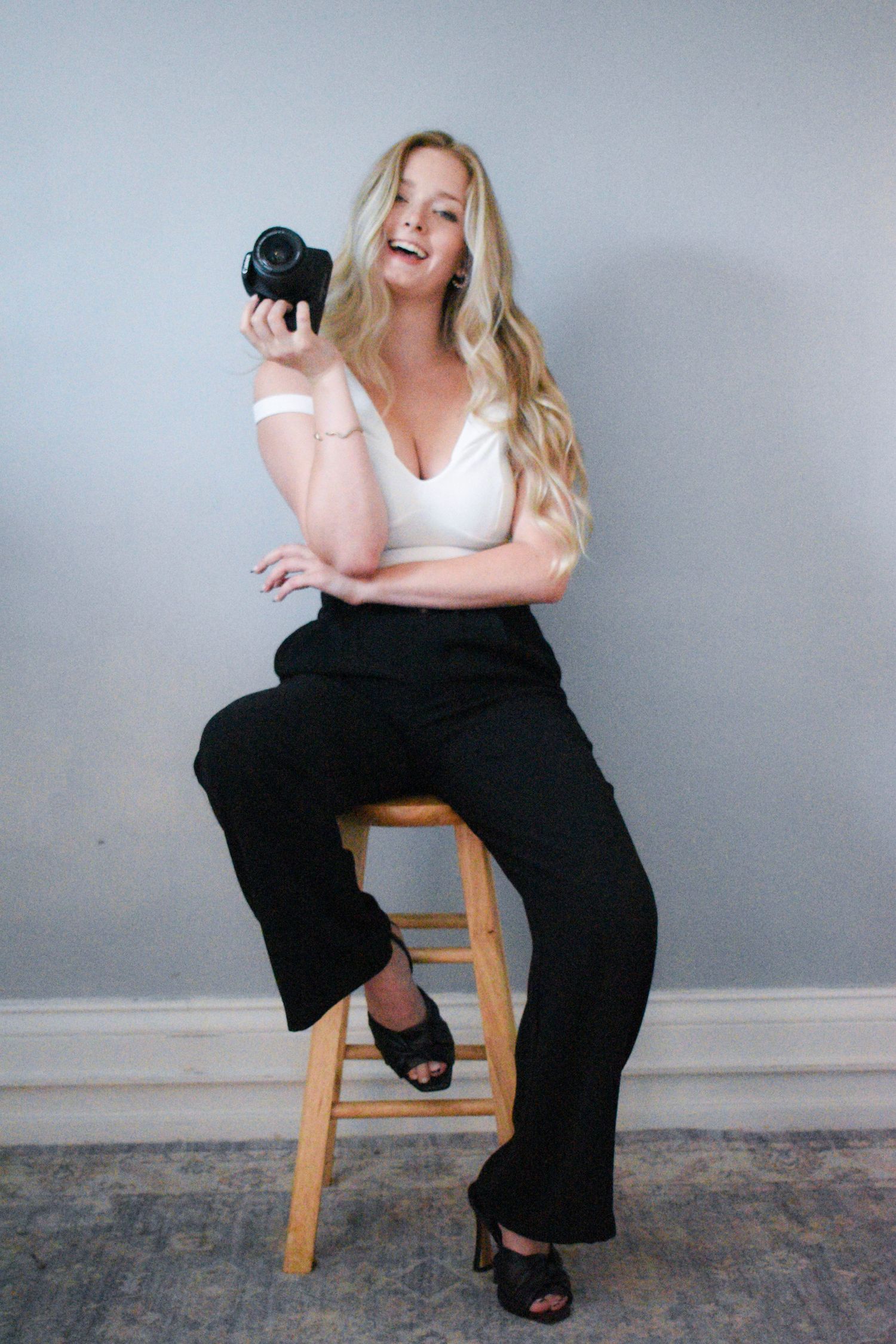 Candid studio shot of person laughing while holding camera on wooden stool.