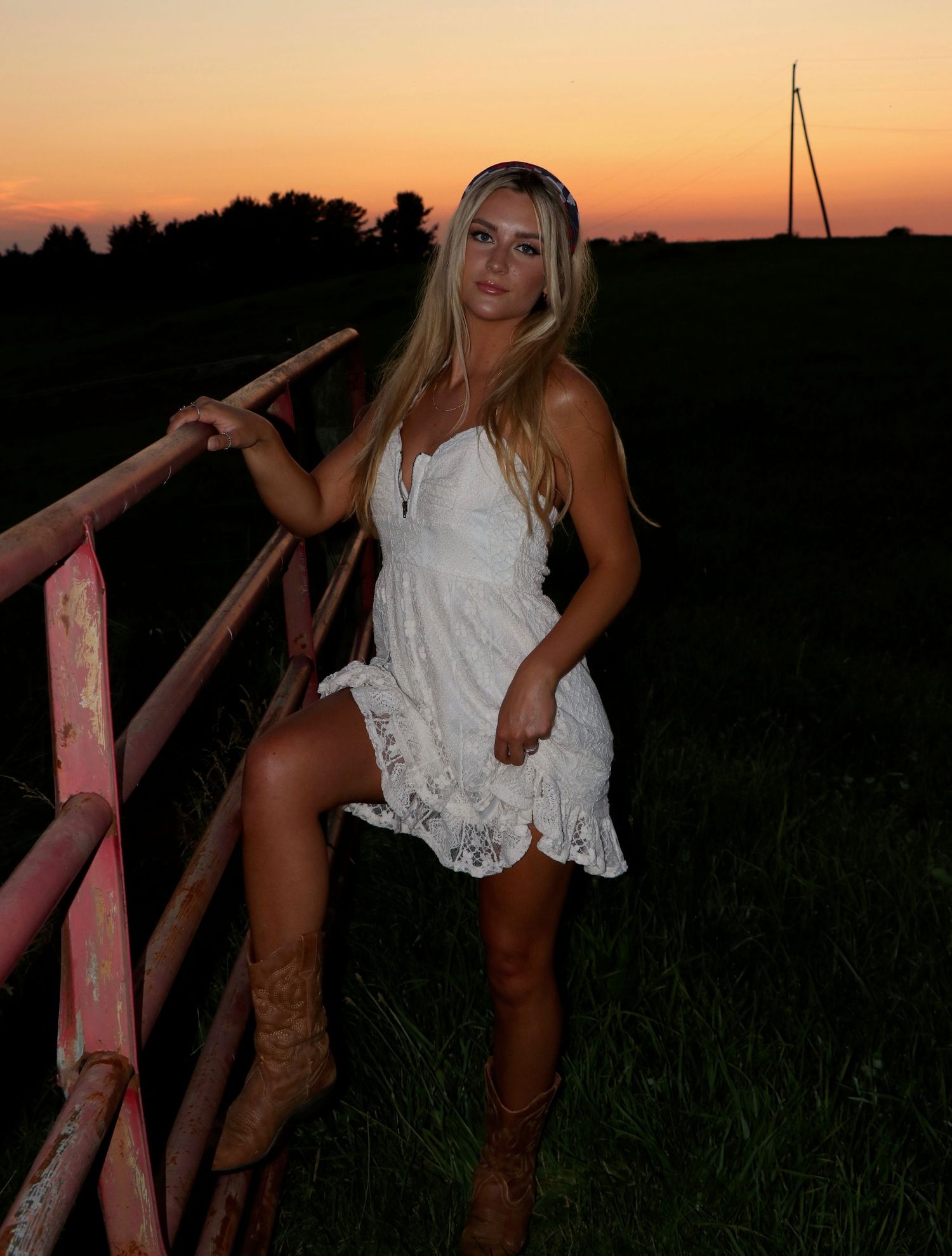Sunset portrait with white lace dress and cowboy boots against rustic wooden railing.