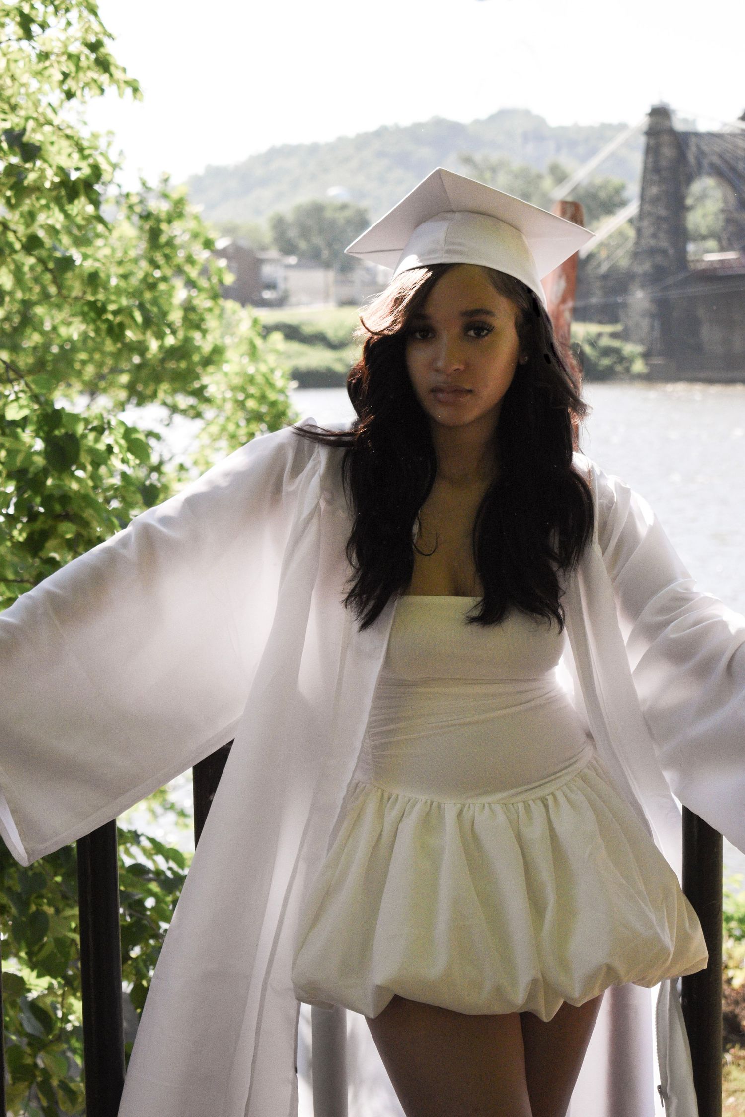 Graduate in white dress and cap poses against scenic mountain backdrop.