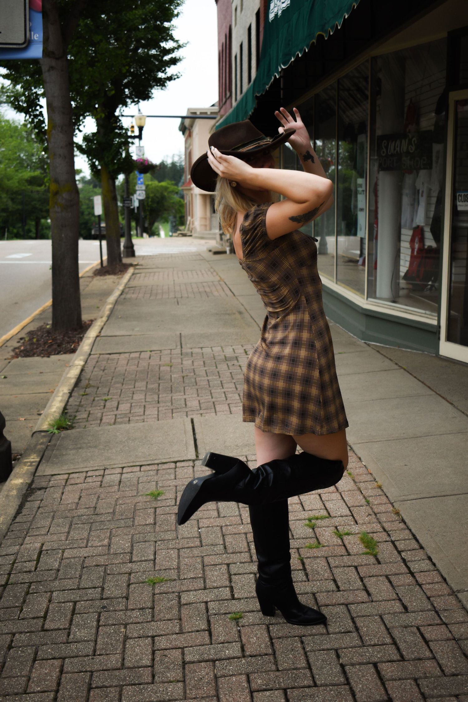 Person in brown plaid dress and black boots poses expressively on urban sidewalk.