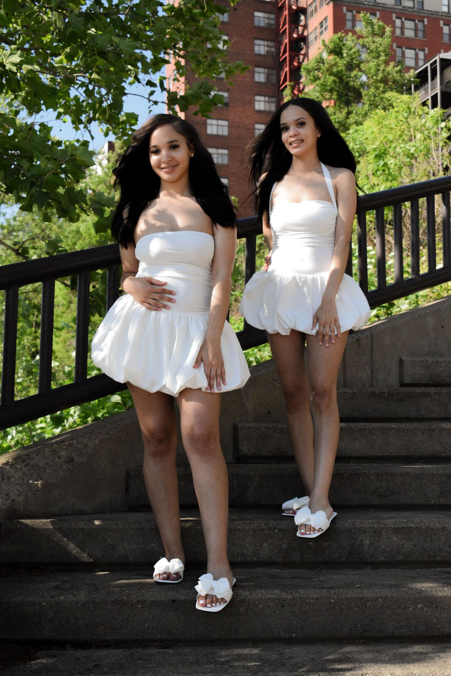 Two women in matching white summer dresses pose on stone steps near brick buildings.