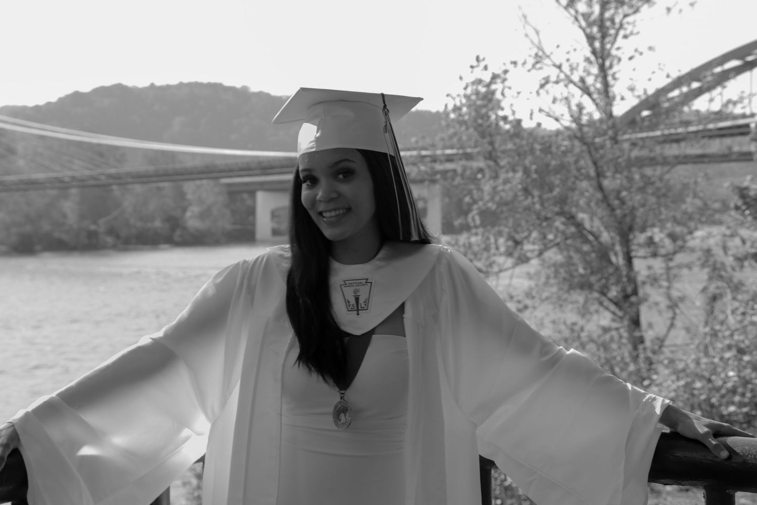 Graduate in white academic regalia poses against scenic mountain backdrop in black and white.