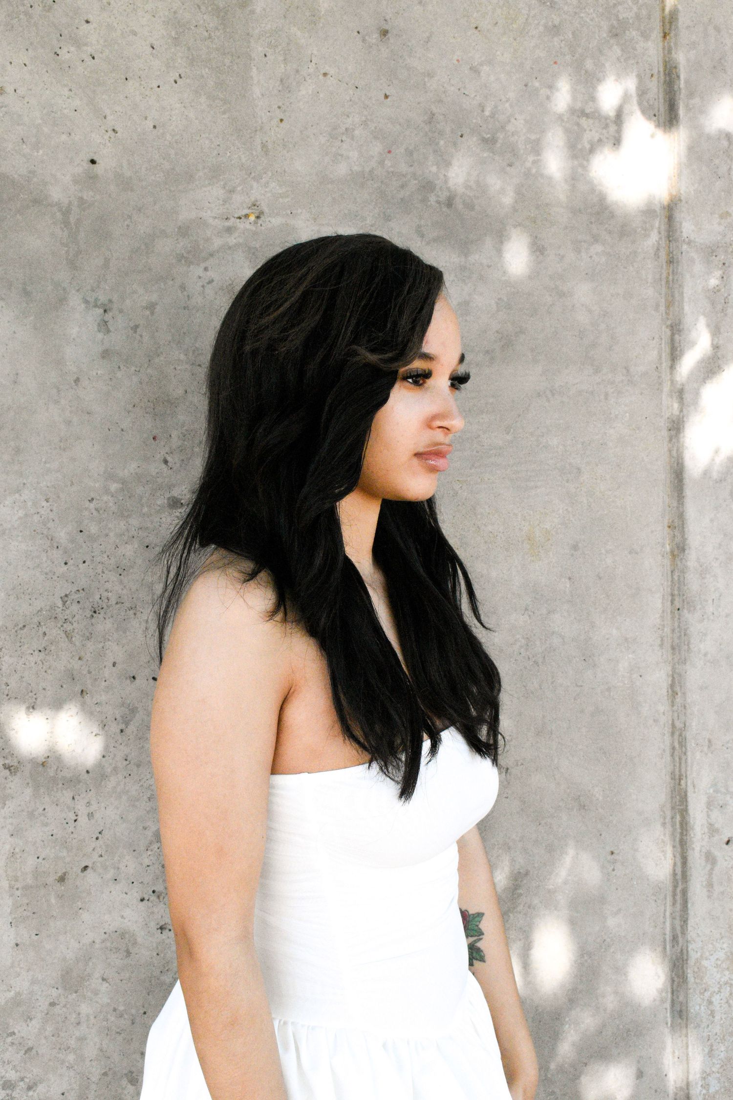 Artistic profile portrait of woman in white dress against concrete wall with dappled sunlight.