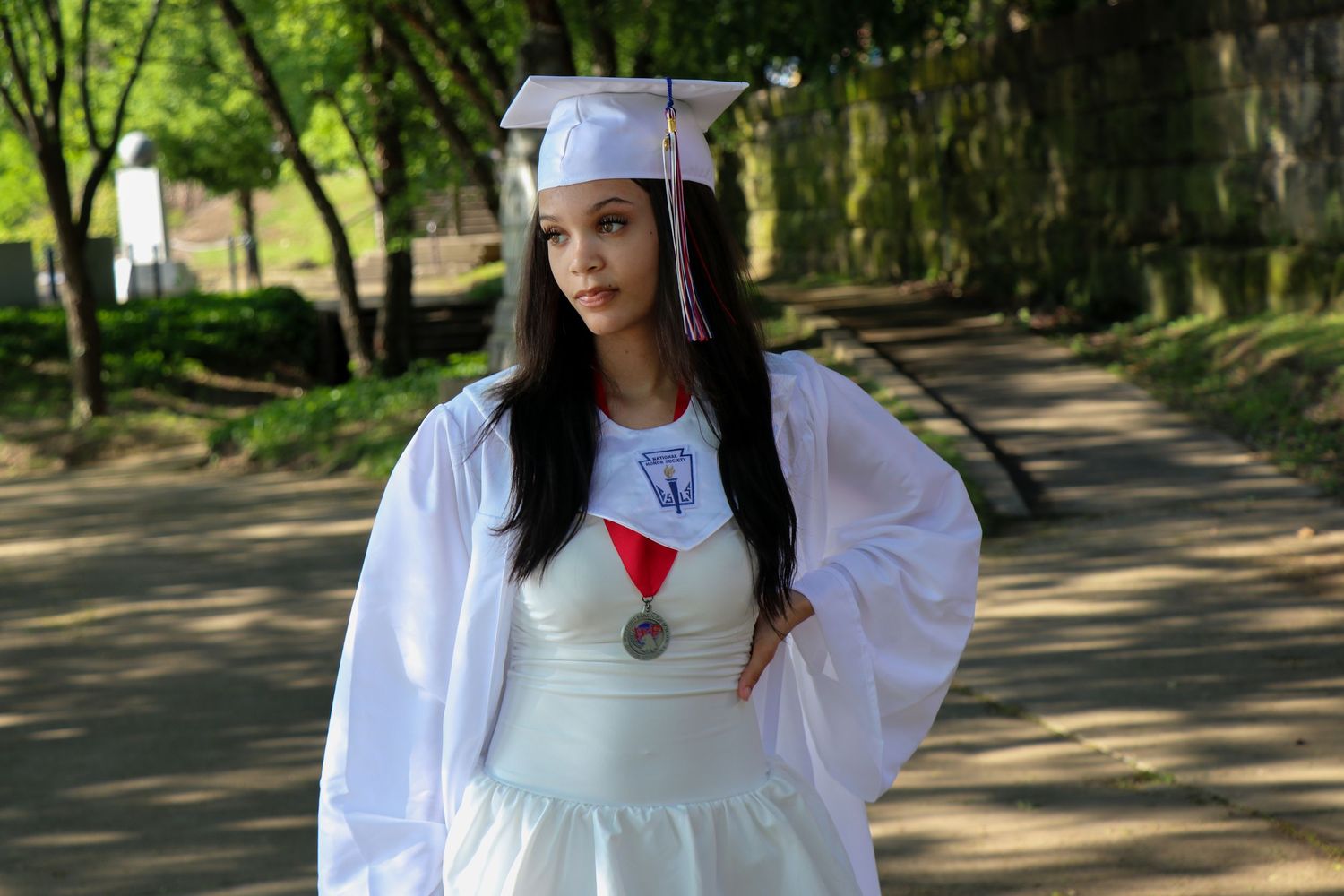 Graduate in white dress and graduation gown poses on outdoor campus pathway.