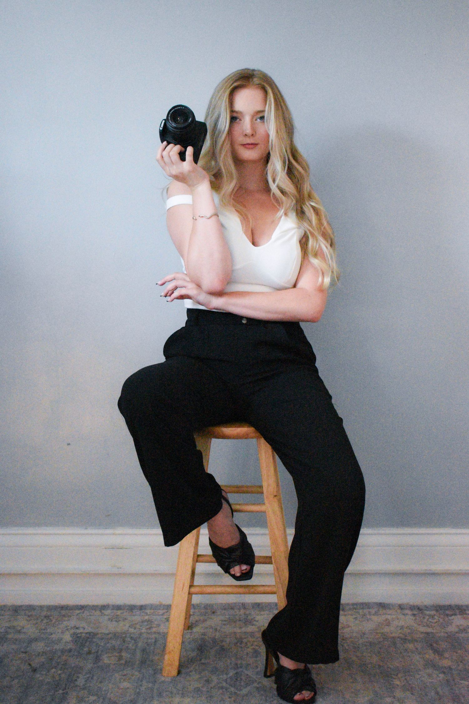 Fashion portrait with person seated on wooden stool holding camera in studio setting.