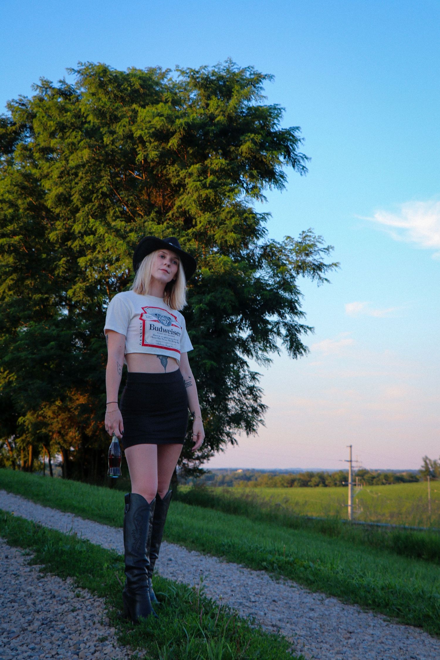 Silhouette on gravel path with evergreen trees and fields in background during sunset.
