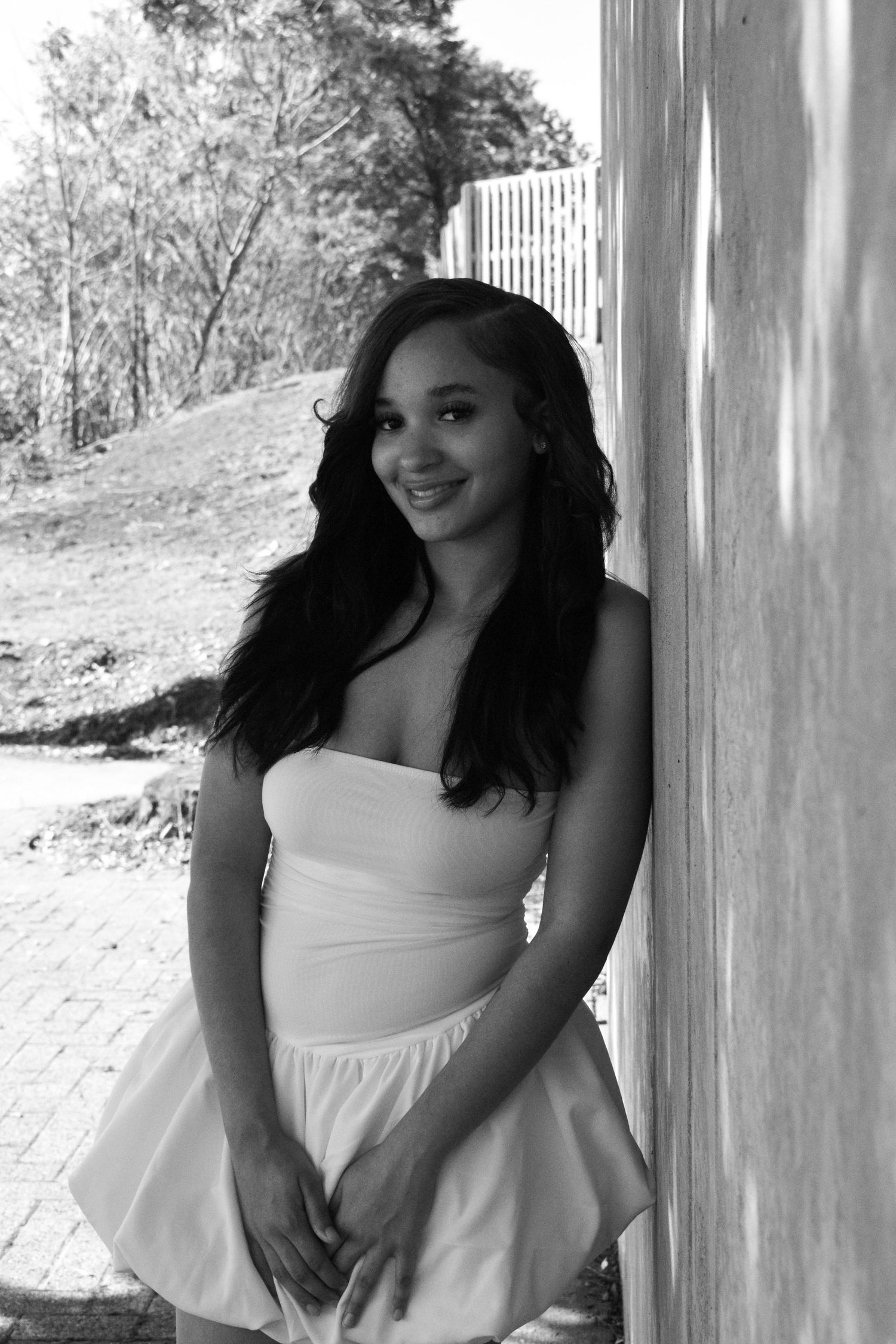 Black and white portrait of woman in strapless white dress standing against wall outdoors.