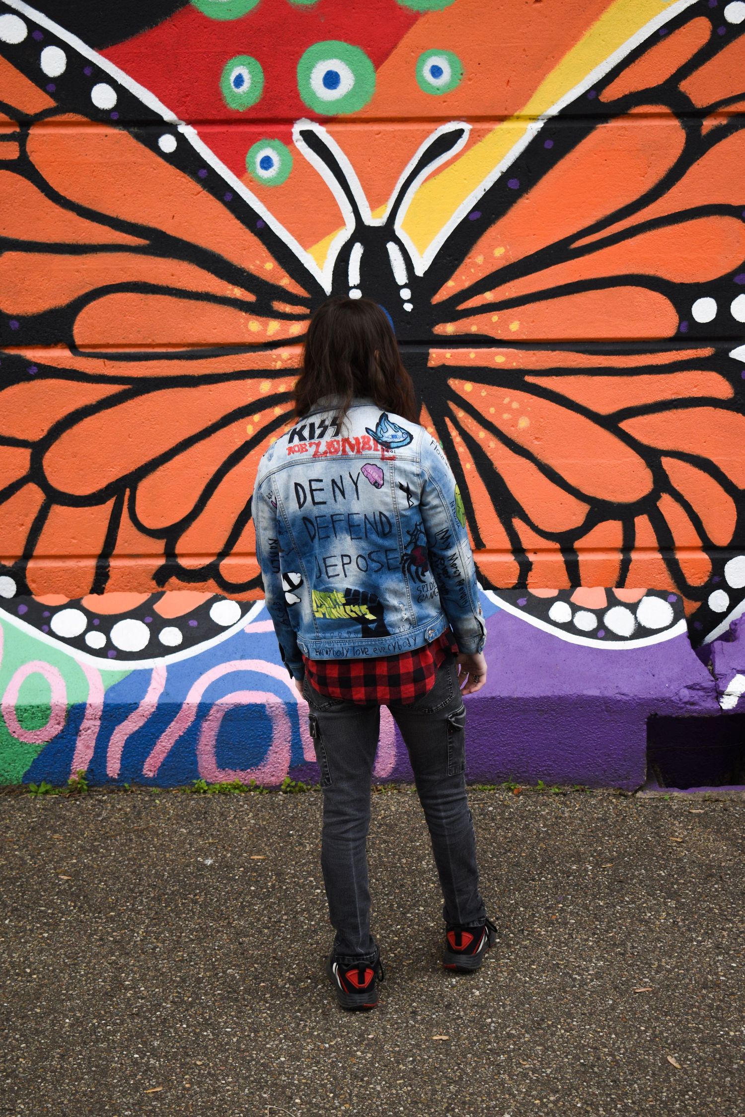 Person wearing denim jacket stands in front of vibrant orange butterfly street art mural.