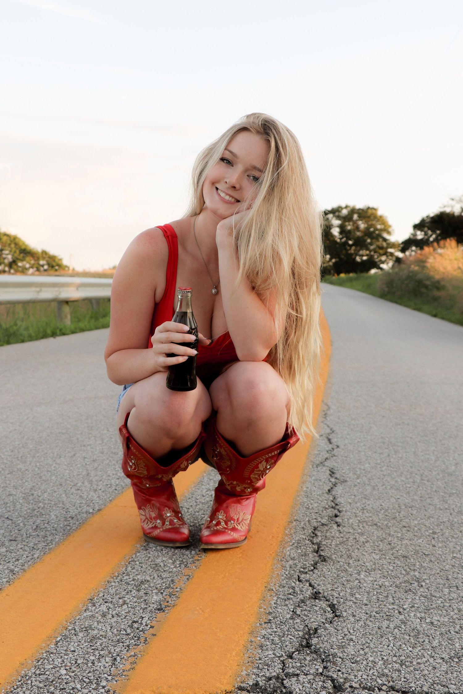 Subject crouching on yellow road lines while holding a beverage at sunset.