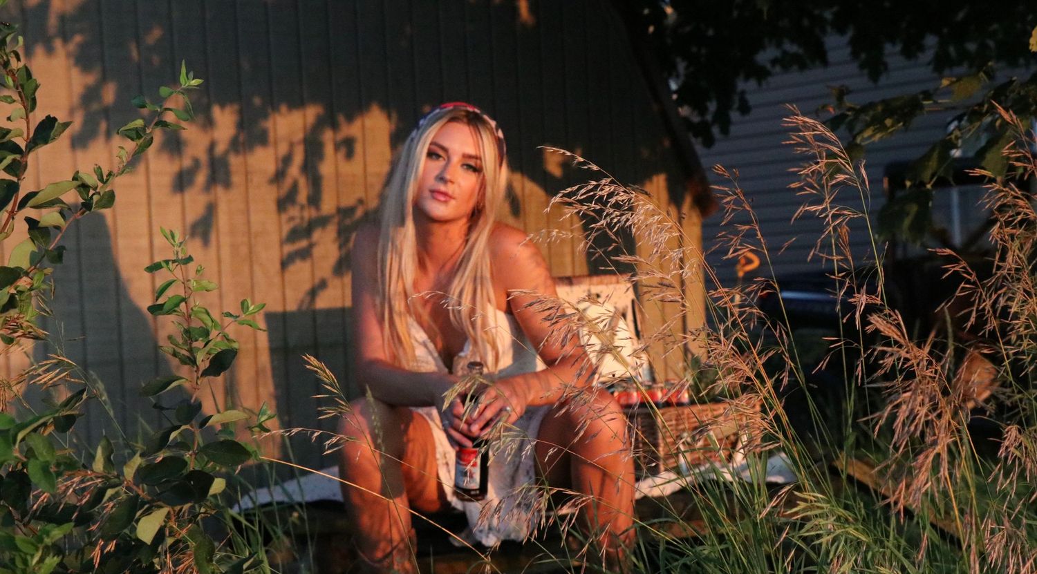 A woman with long hair sits in a garden at golden hour while surrounded by wild grasses and plants.