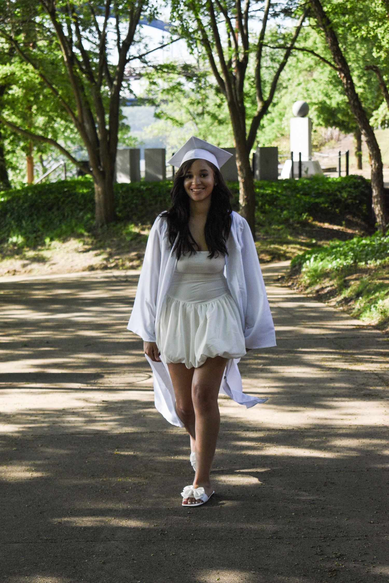Graduate in white dress and cap walking down a tree-lined path on graduation day.