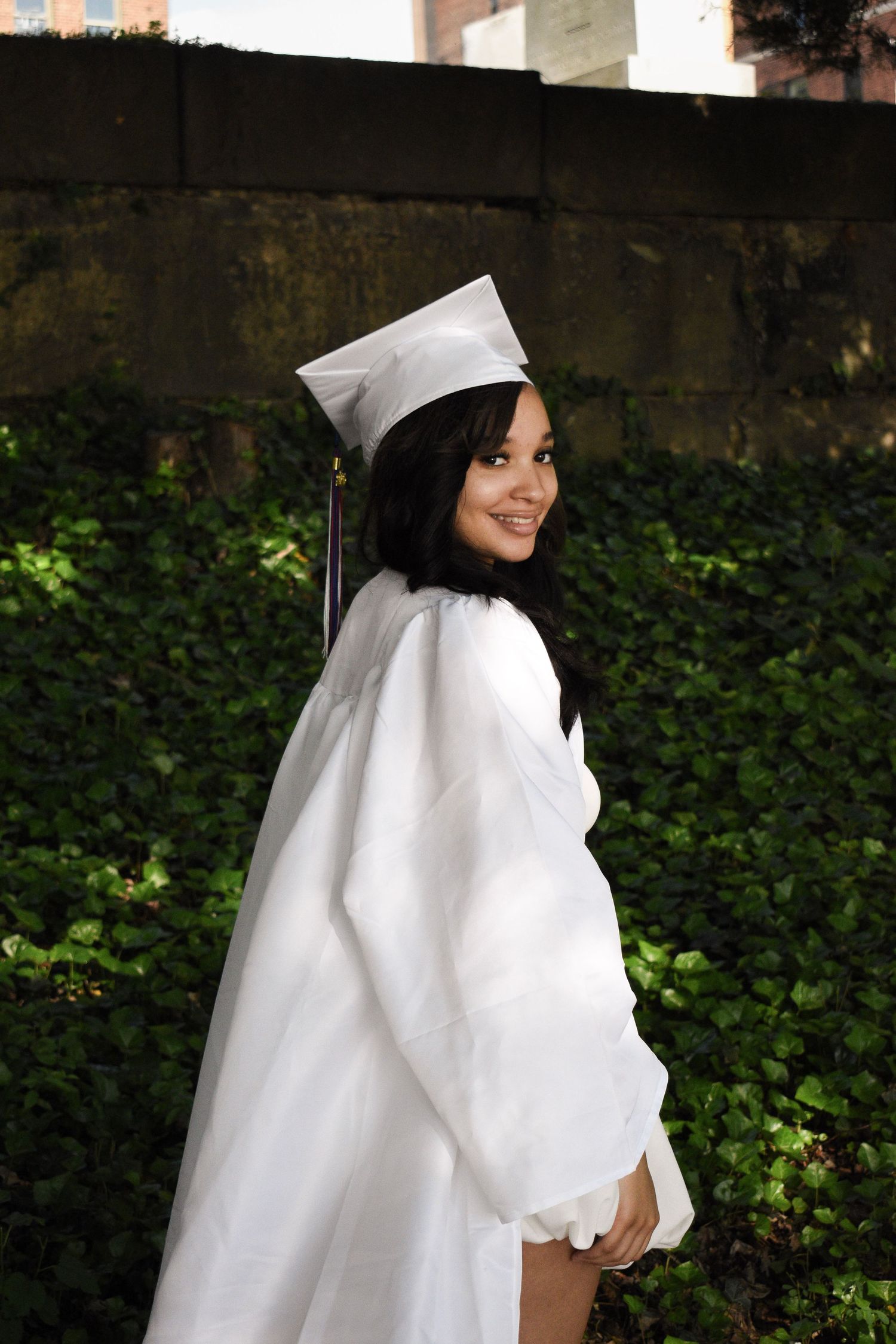 Graduate in white cap and gown poses against an ivy-covered wall outdoors.