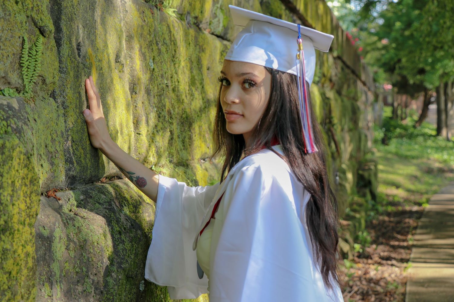 Graduate in white cap and gown poses against mossy stone wall in nature setting.