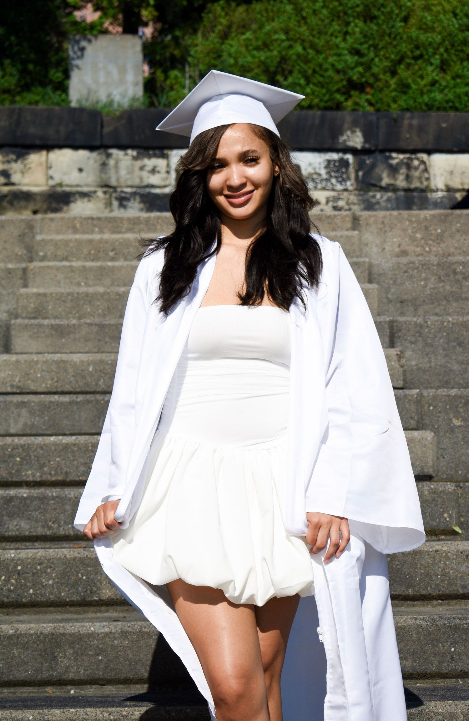 Graduate in white dress and cap poses on stone steps during sunny graduation photo shoot.