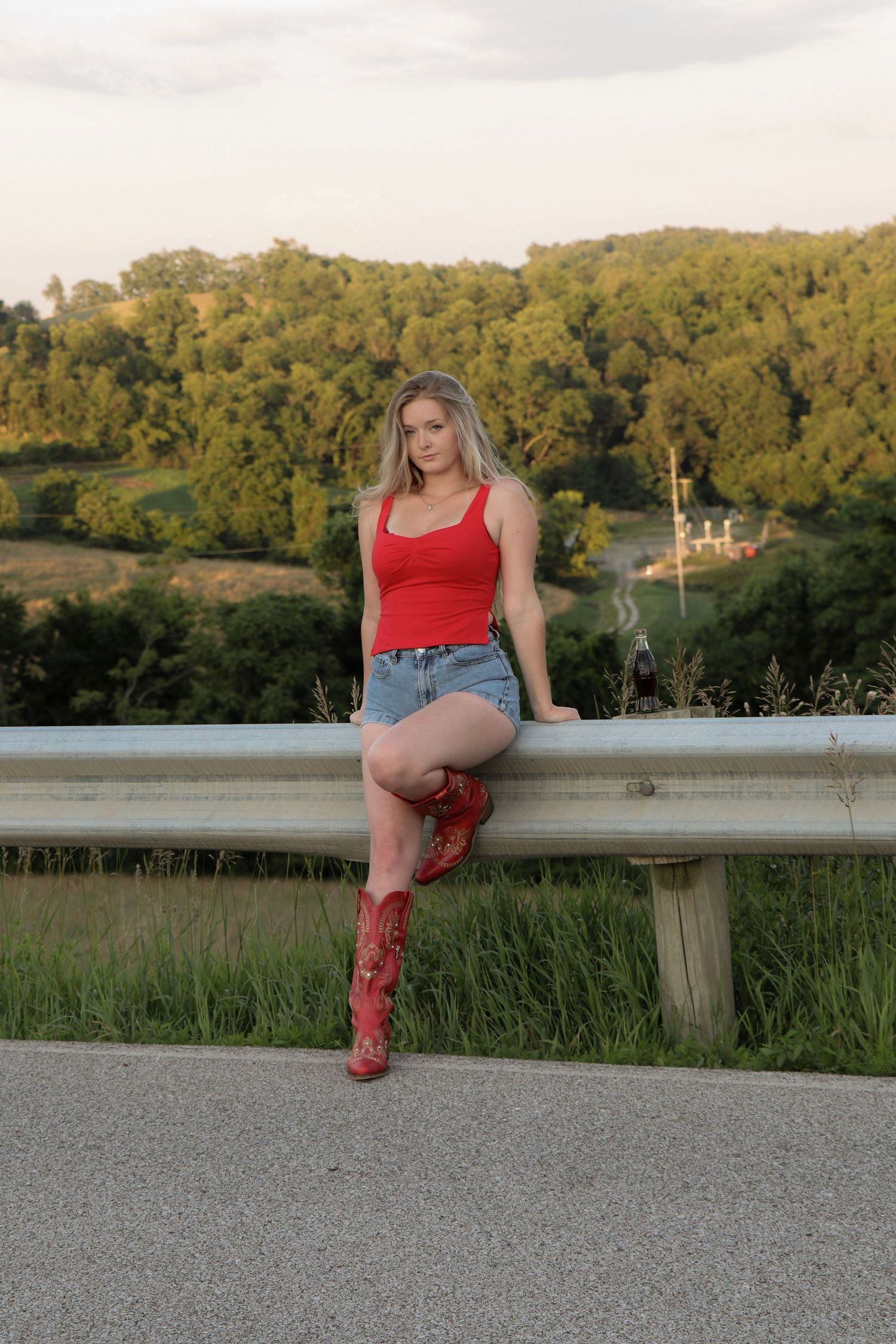 Person in red tank top and denim sitting on a guardrail overlooking green hills at sunset.