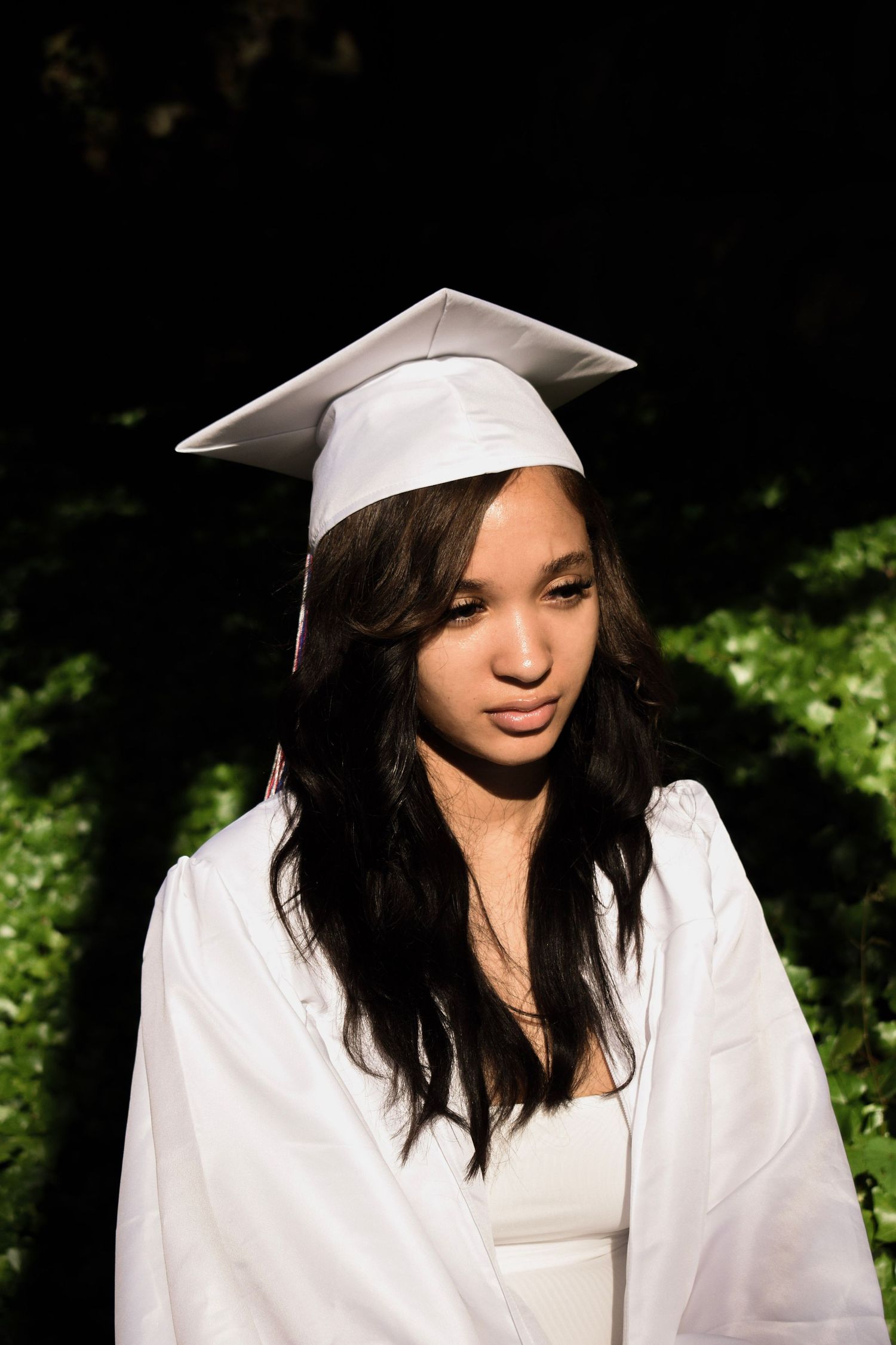 Graduate in white cap and gown photographed in dramatic lighting outdoors.
