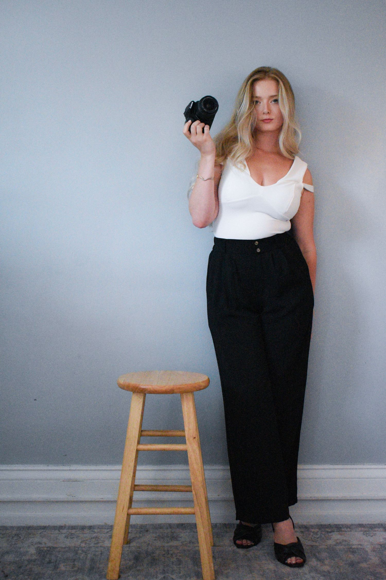 Person in white top and black pants poses with camera next to wooden stool against grey wall.