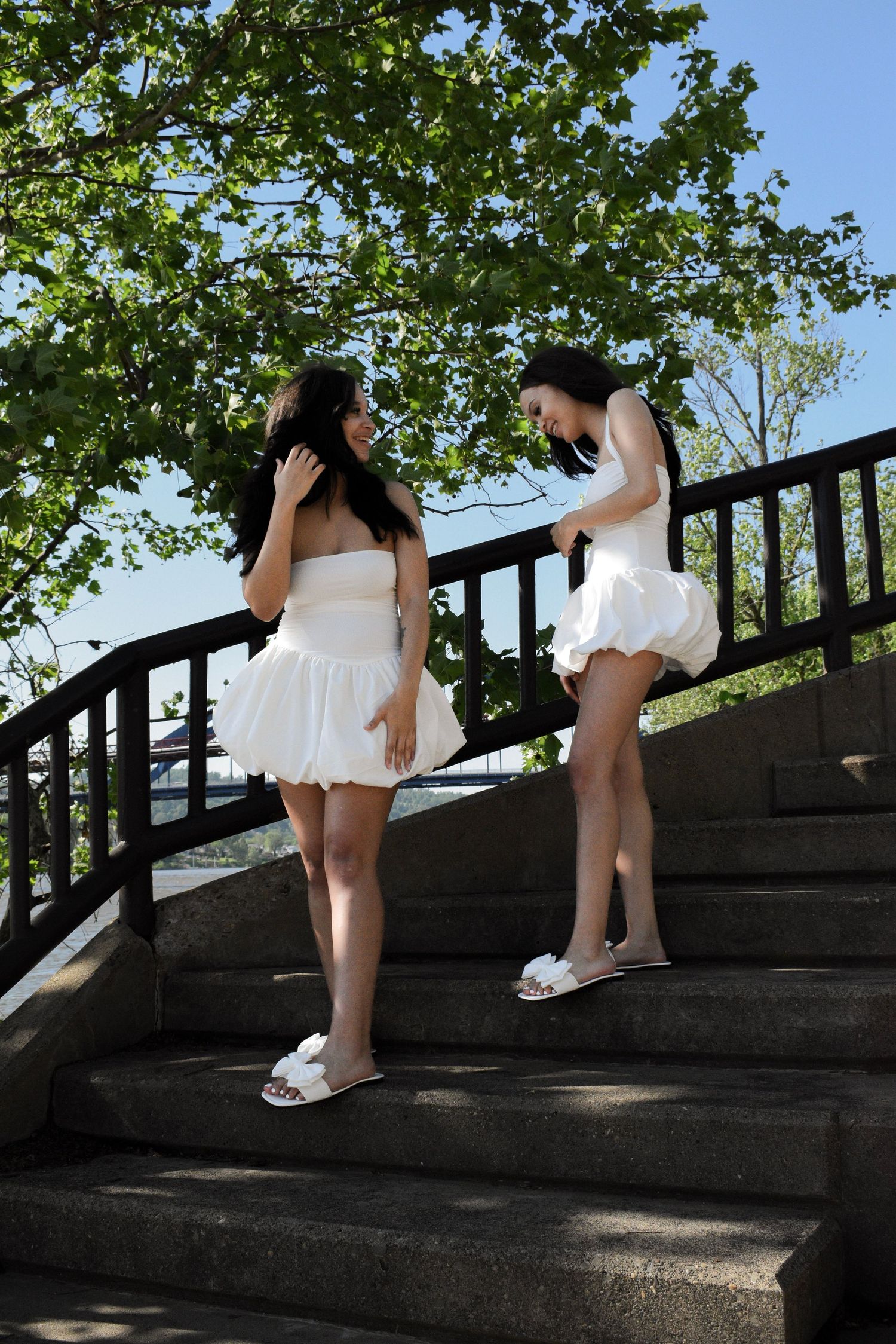 Two friends in white dresses and sandals chatting on outdoor stone staircase under leafy trees.