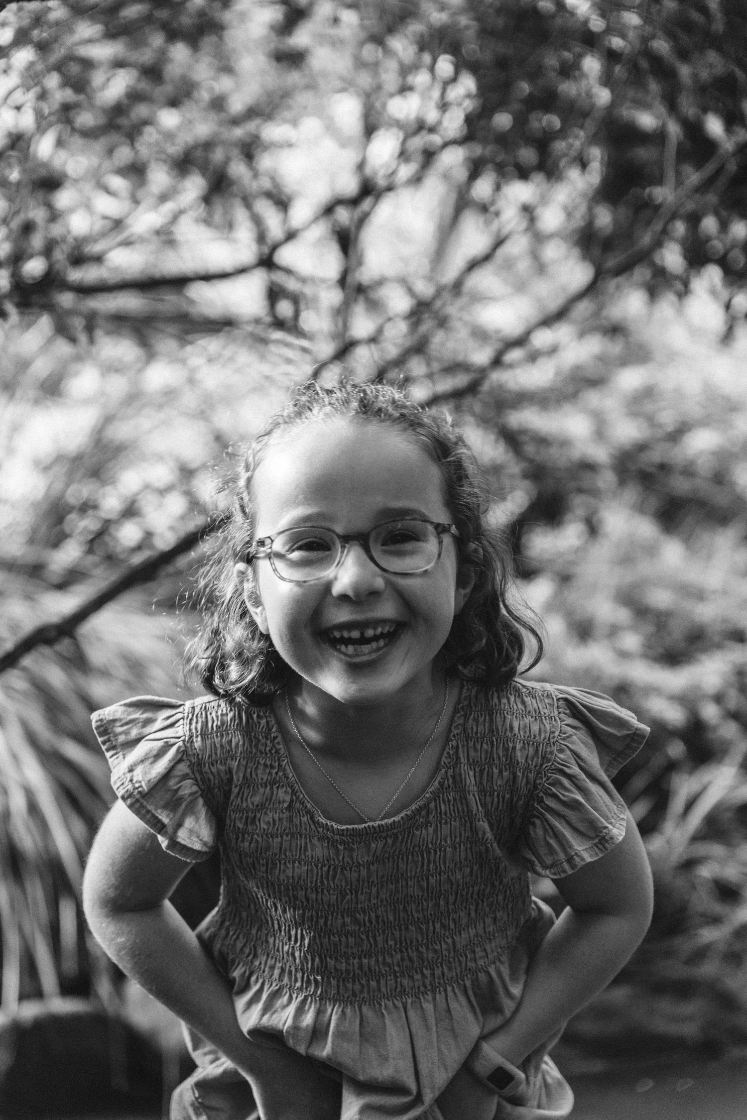 Black and white portrait of a joyful child wearing glasses in a garden setting. Illawarra