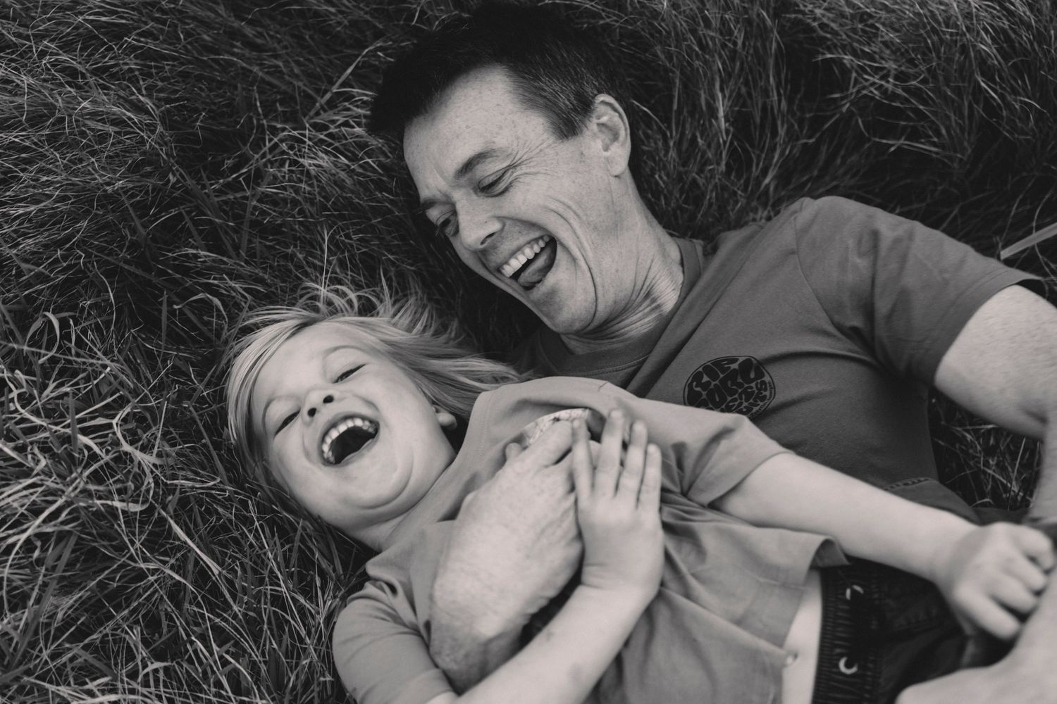 Candid black and white photo of playful laughter while laying in tall grass. Gerringong
