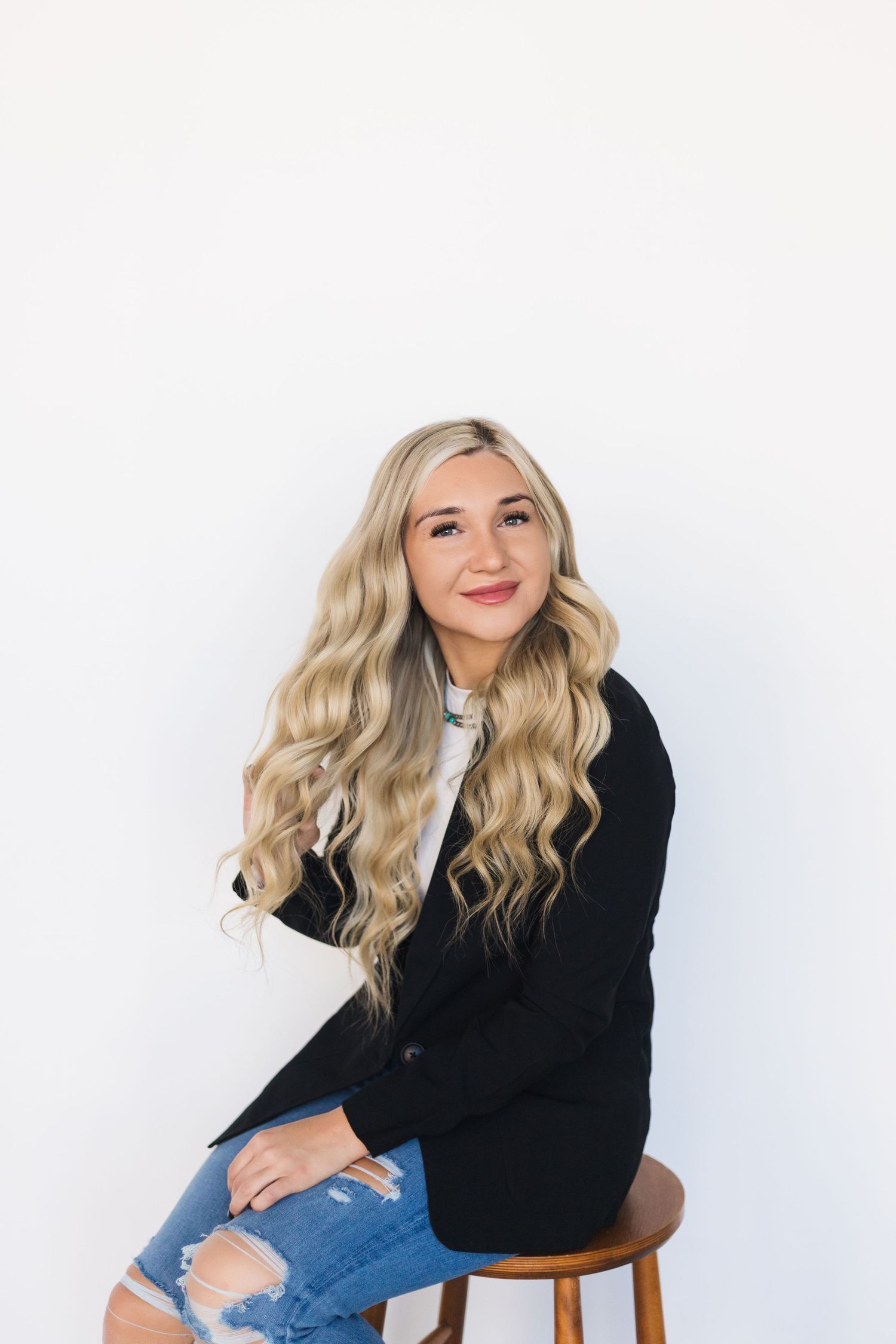 Studio portrait in black cardigan and distressed jeans seated on wooden stool.