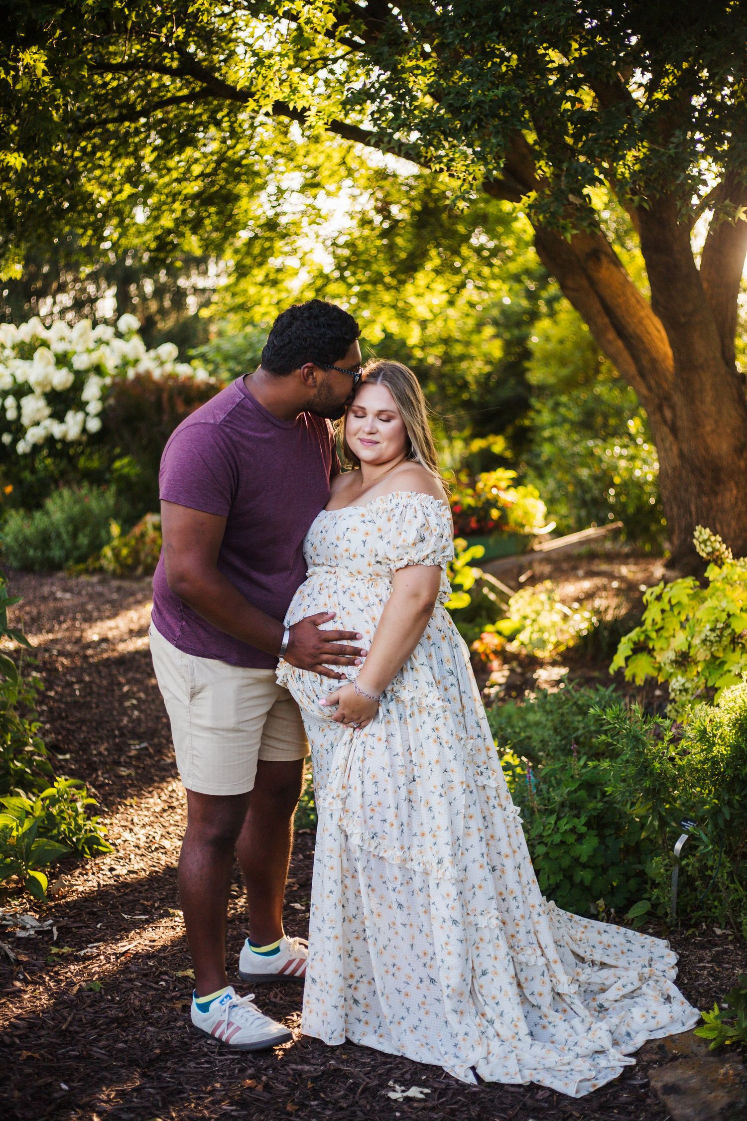 A couple poses for a romantic maternity photo in a sunlit garden setting with a flowing white lace dress.