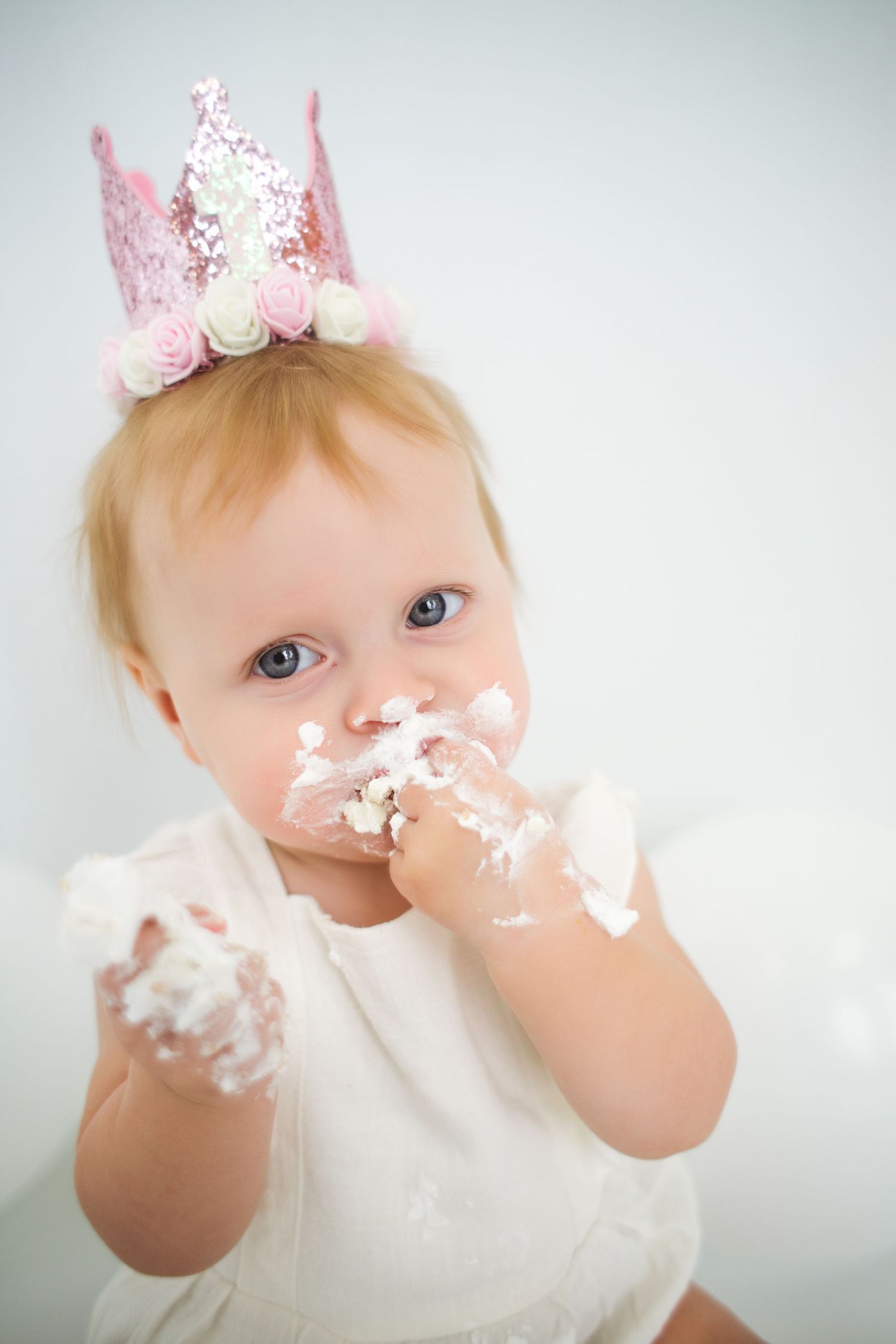 A baby wearing a pink crown enjoys smashing and eating birthday cake with frosting all over their face.