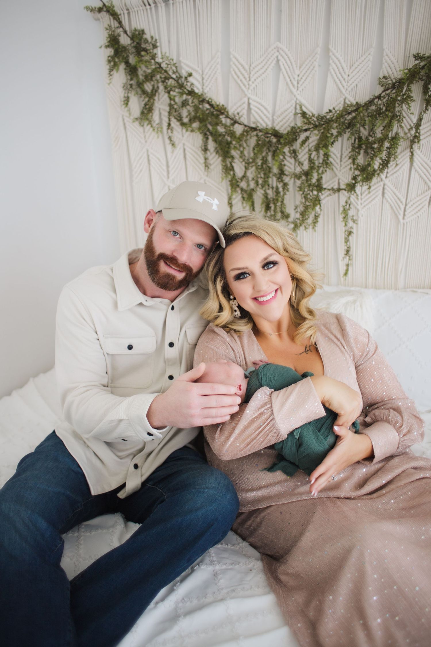 A couple sits together on a bed beneath a pine garland and macrame wall hanging in a cozy indoor setting.
