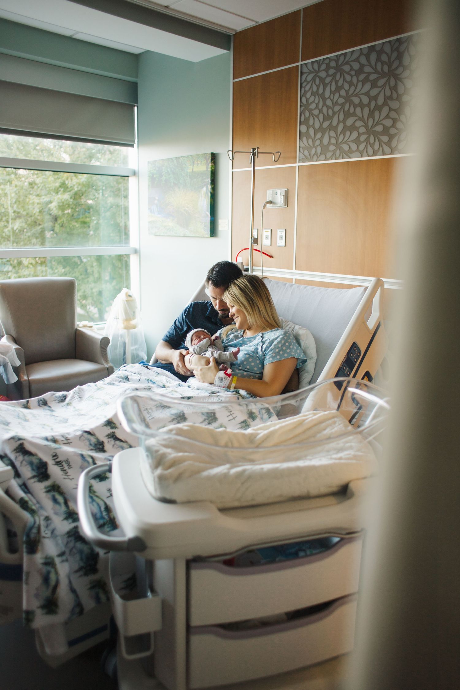 A private hospital room with mint green walls, wood accents, and a medical bed near a large window with natural lighting.