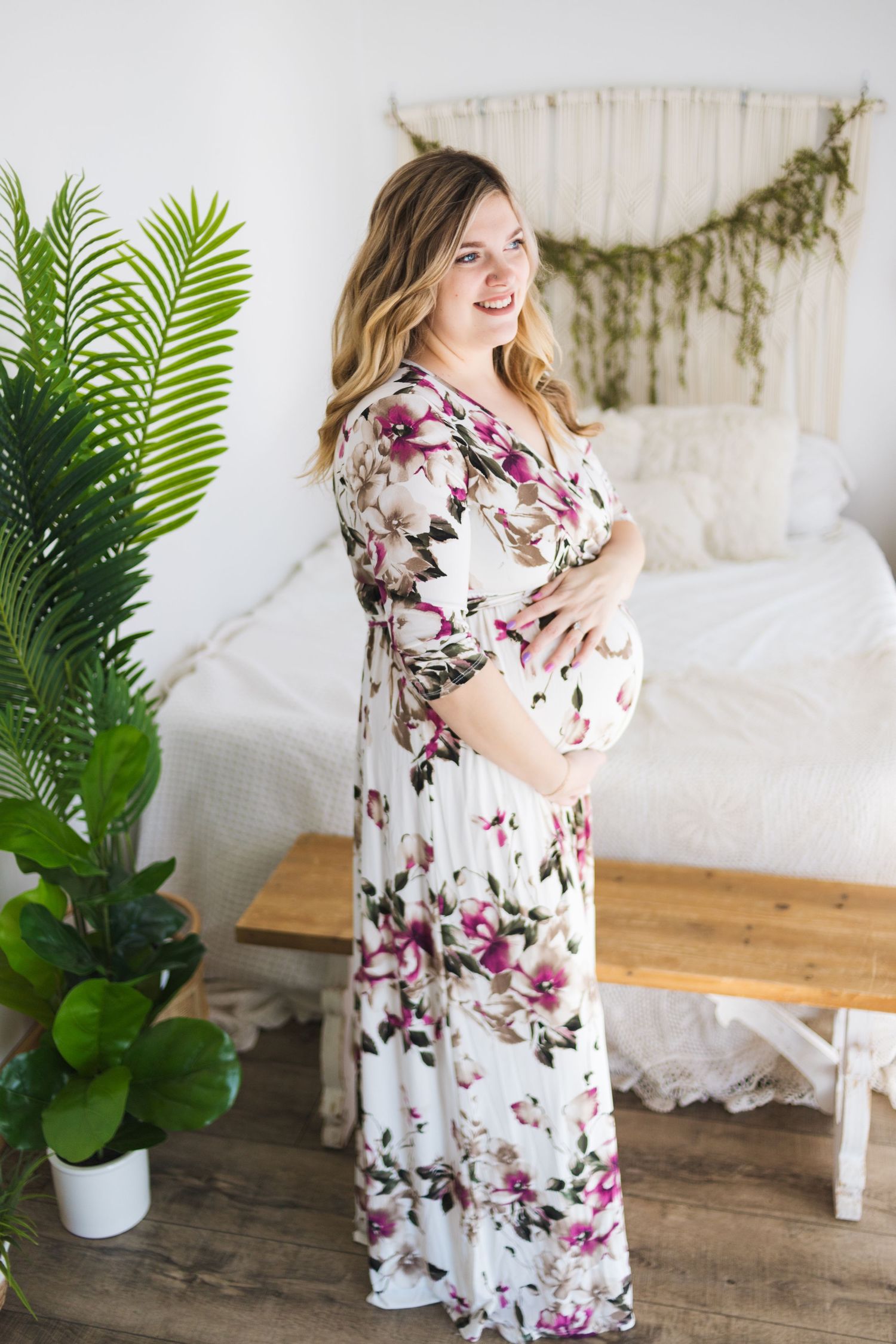 An expectant mother in a floral maxi dress poses against a white backdrop with tropical plants.