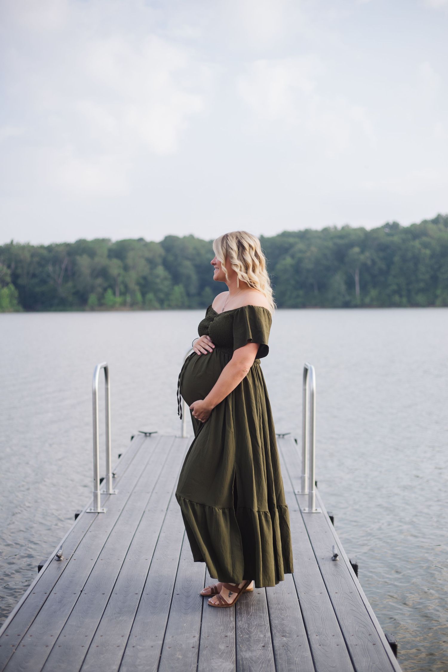 A person in a dark green dress sits contemplatively on a wooden dock overlooking a serene lake surrounded by trees.