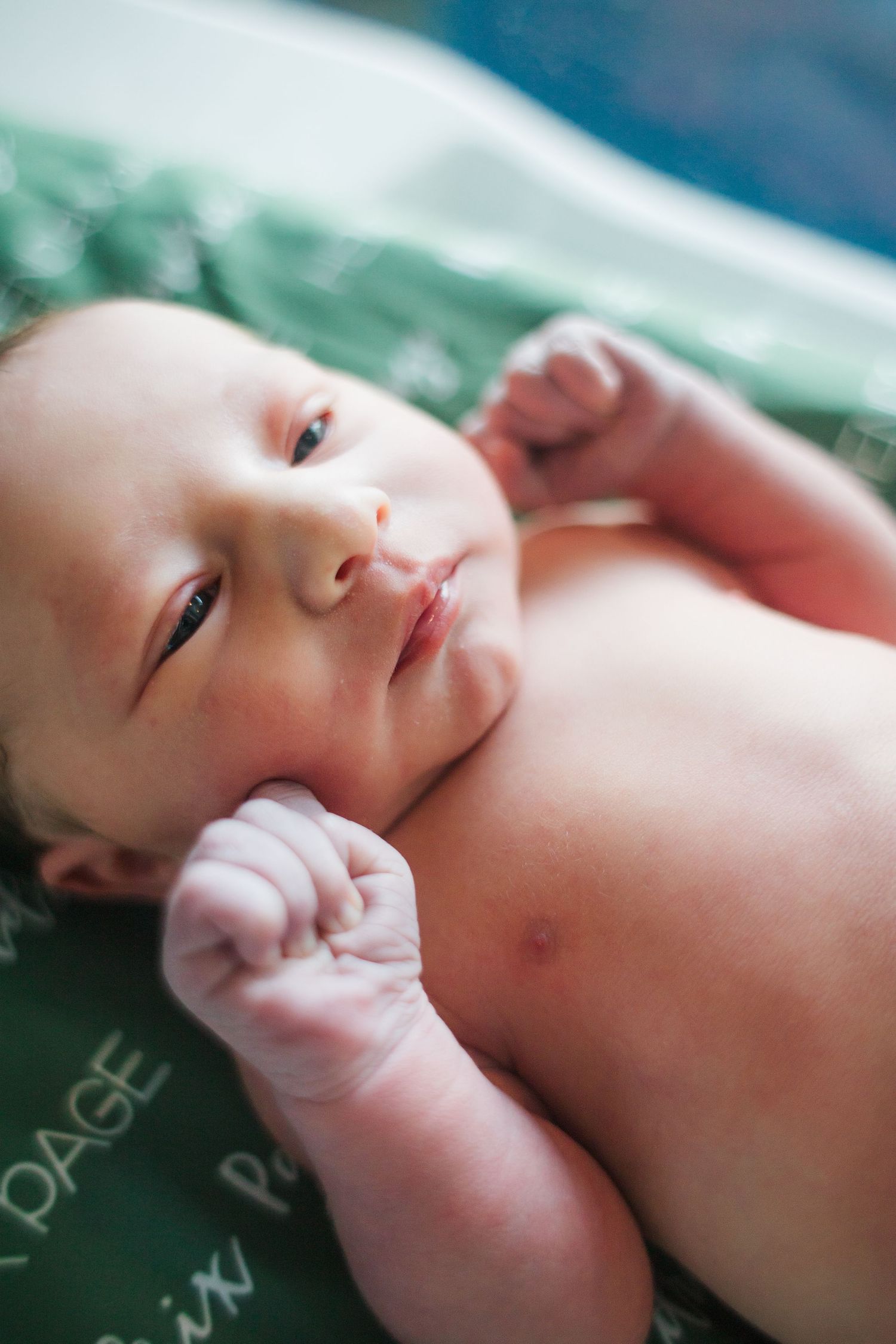 A newborn baby lies on green fabric with tiny fists curled up near their face.