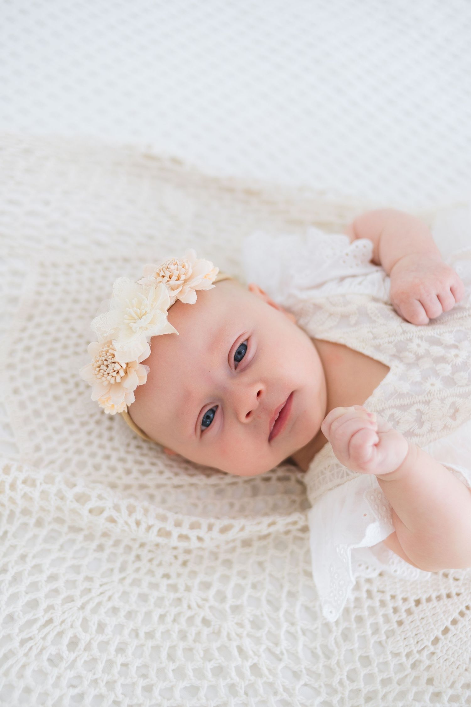A baby lies on white lace fabric wearing a delicate cream flower headband in a series of soft, dreamy portraits.