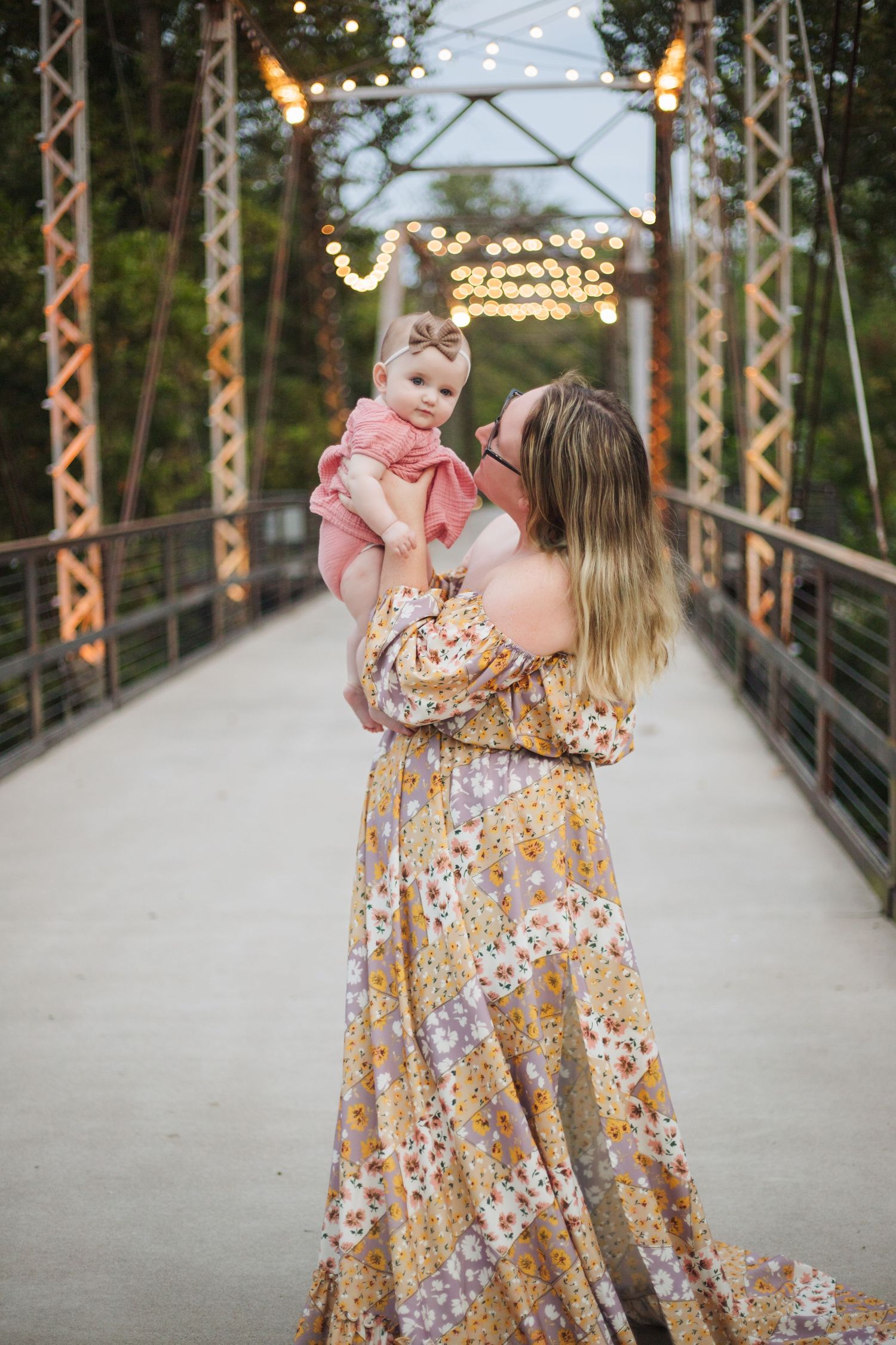 Parent and child share a joyful moment on a bridge with string lights and metal railings at dusk.