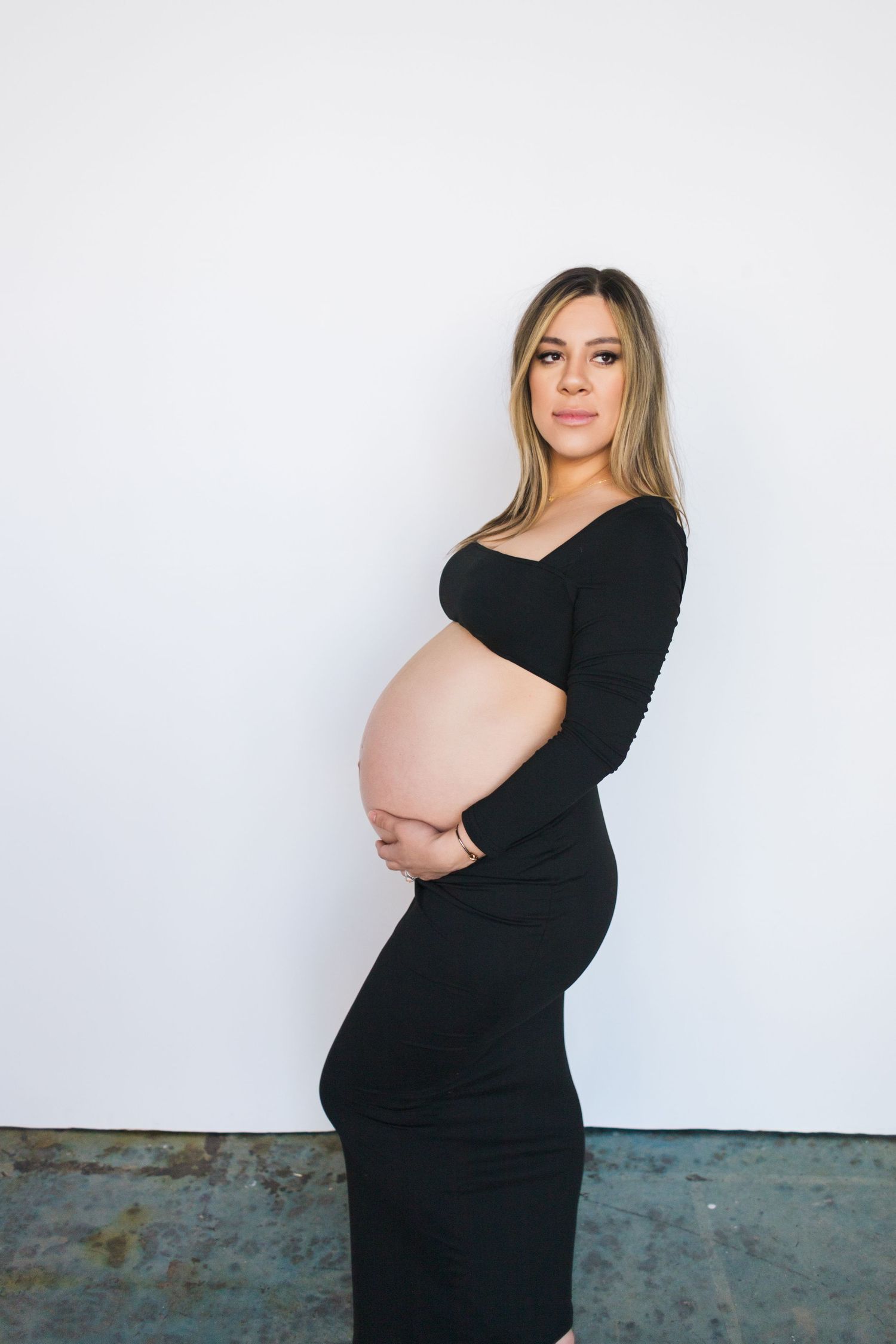 A pregnant woman in a sleek black dress poses against a white wall in a minimalist maternity photo.