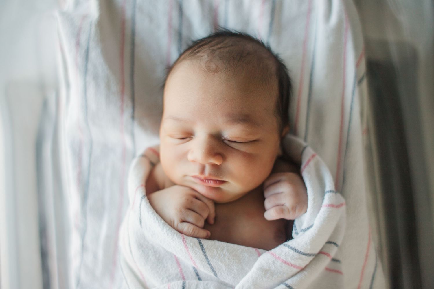 A newborn baby sleeps peacefully wrapped in white blankets on a soft white surface.