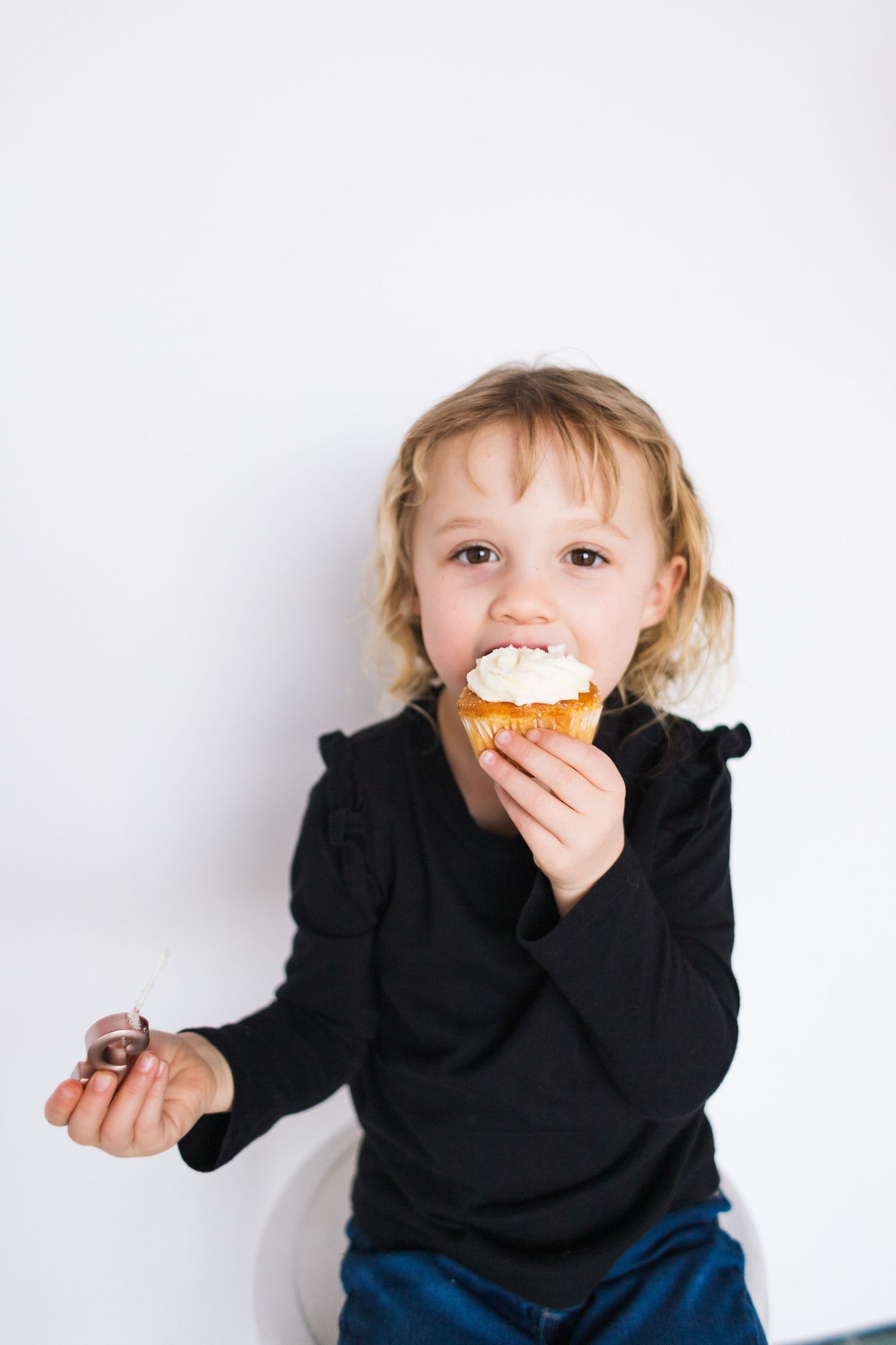 Child in black sweater enjoys a cupcake against white background.