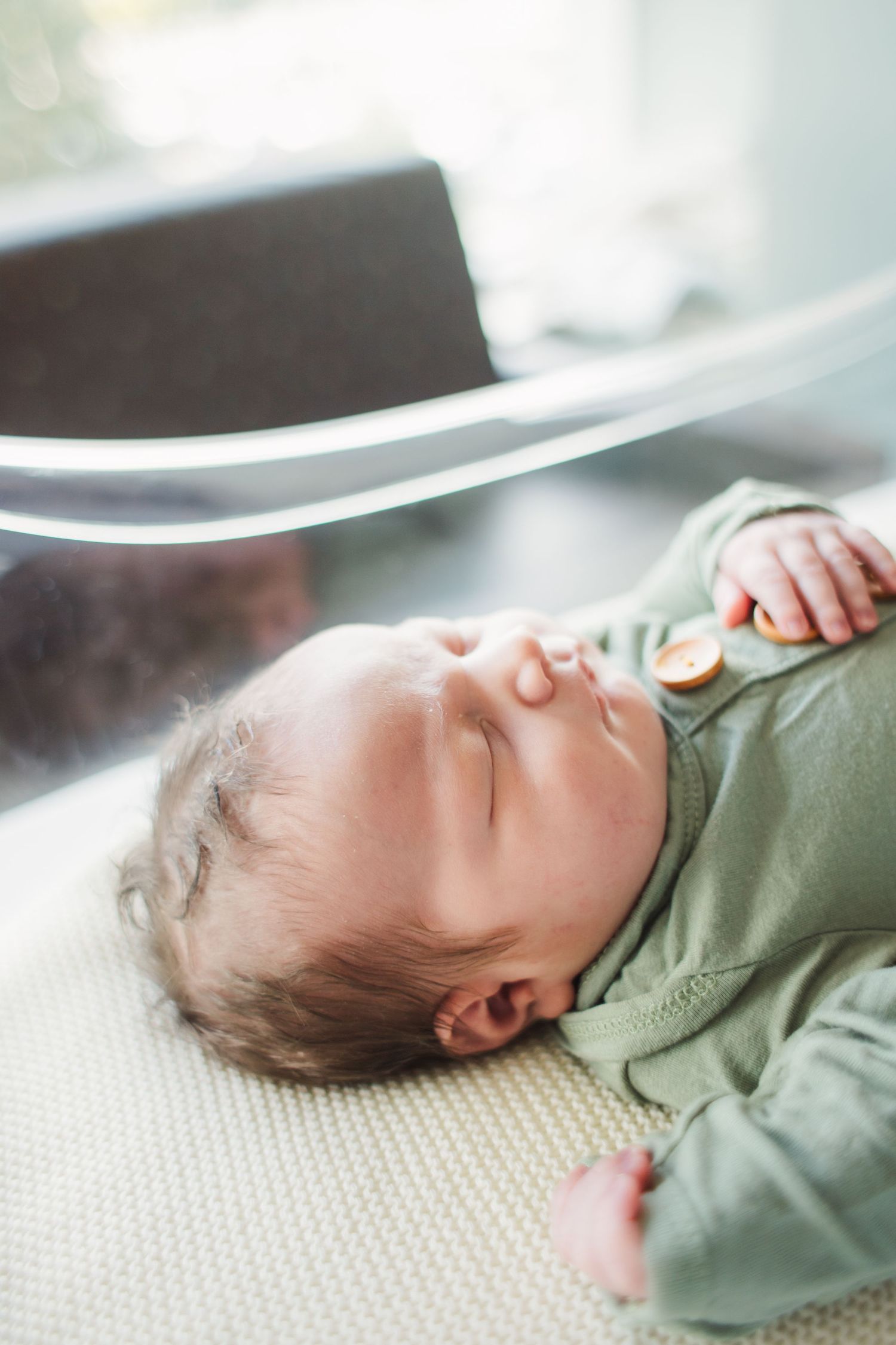 A sleeping infant wearing a sage green outfit lies peacefully on a textured white surface in soft lighting.