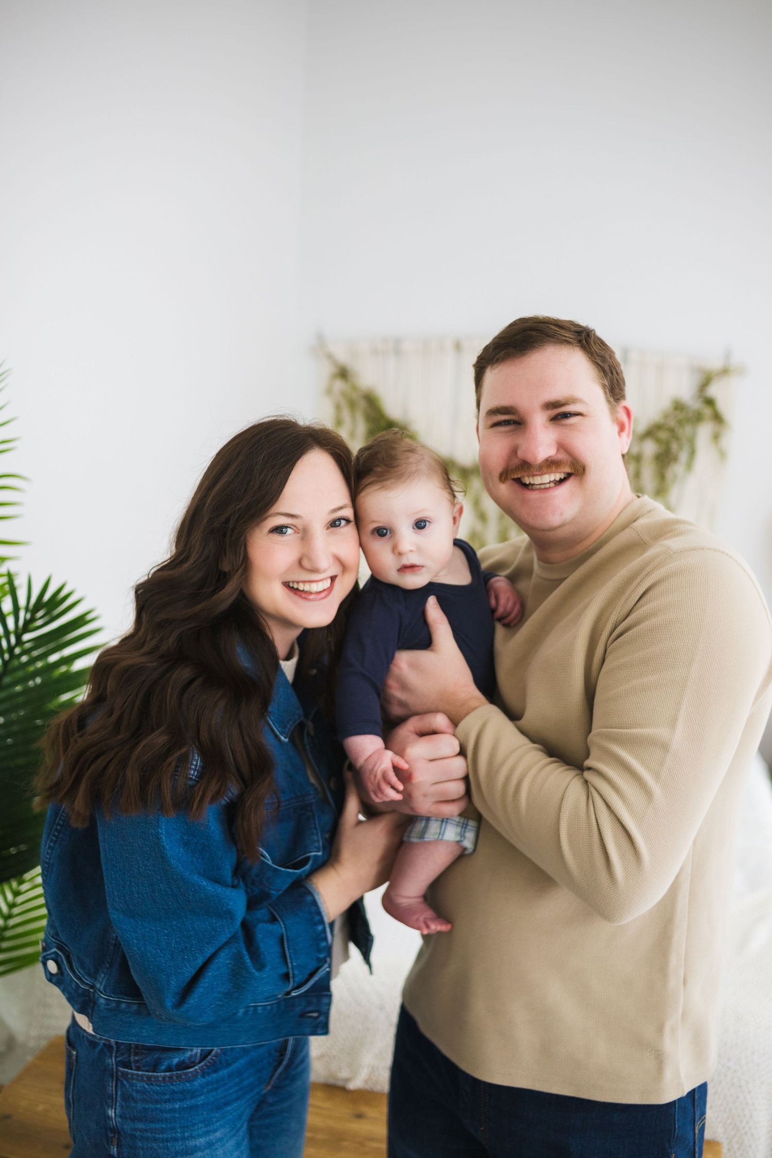Joyful indoor family photoshoot with parents and baby against white wall with greenery.