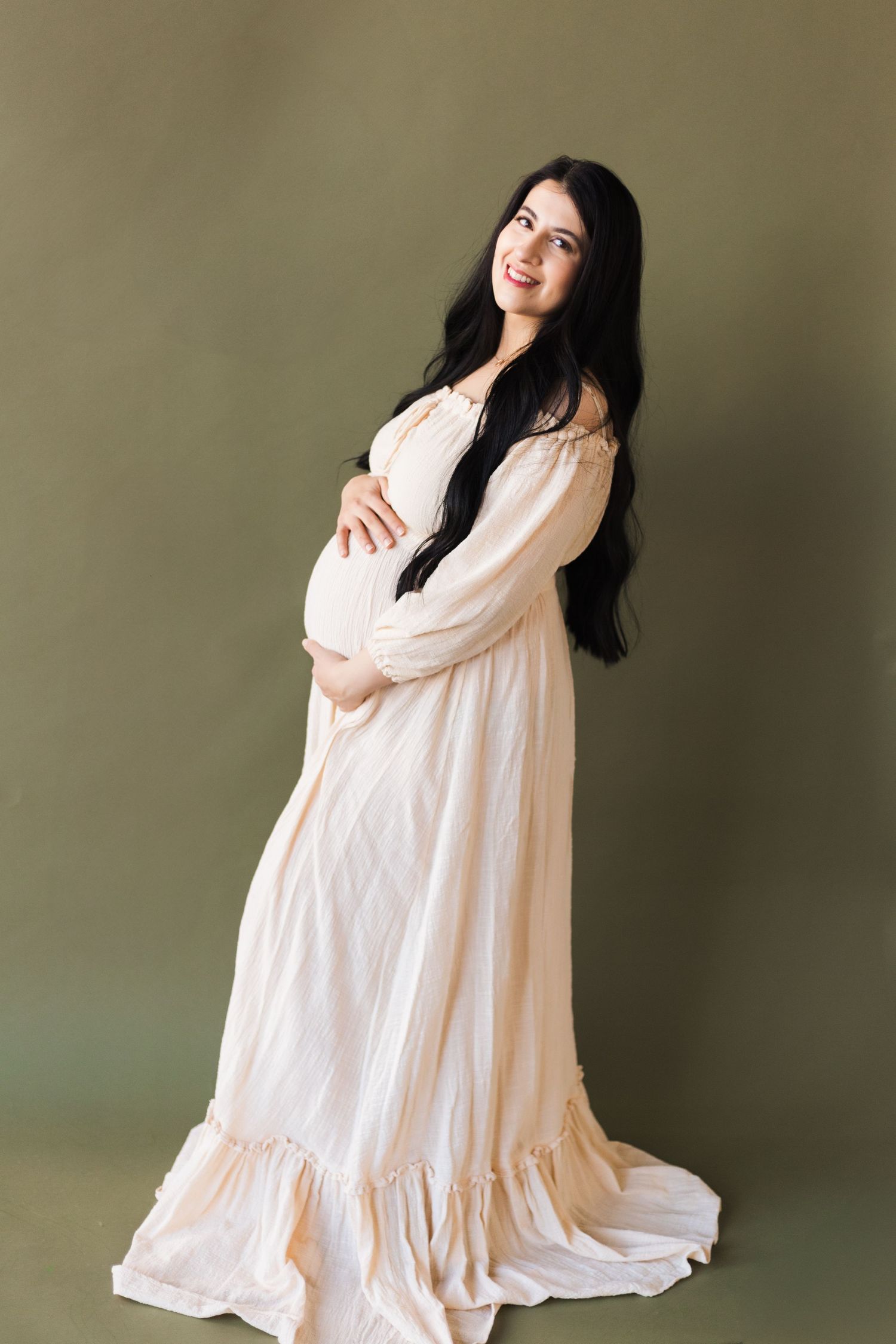 Elegant maternity photo of woman in flowing cream dress against neutral backdrop.