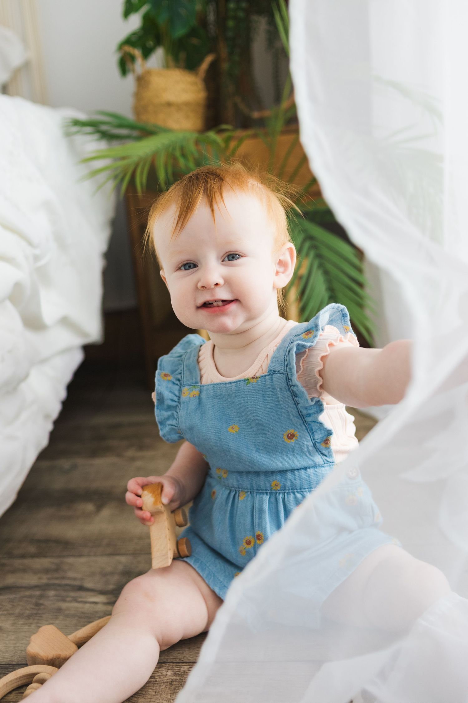 Cheerful baby in blue romper plays on floor near white curtains and tropical plants.