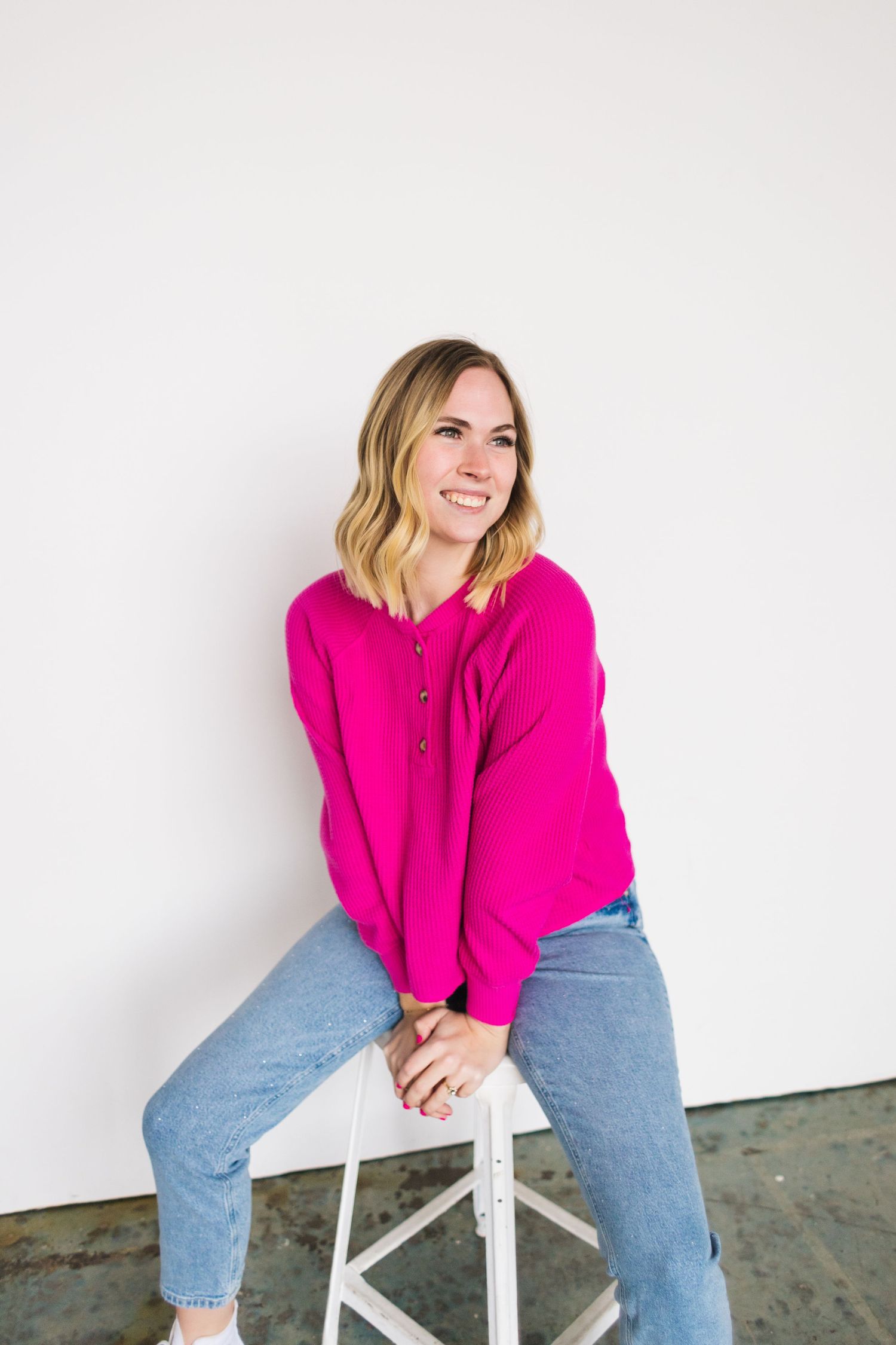 Person in bright pink sweater and jeans sits casually on white stool against plain background.