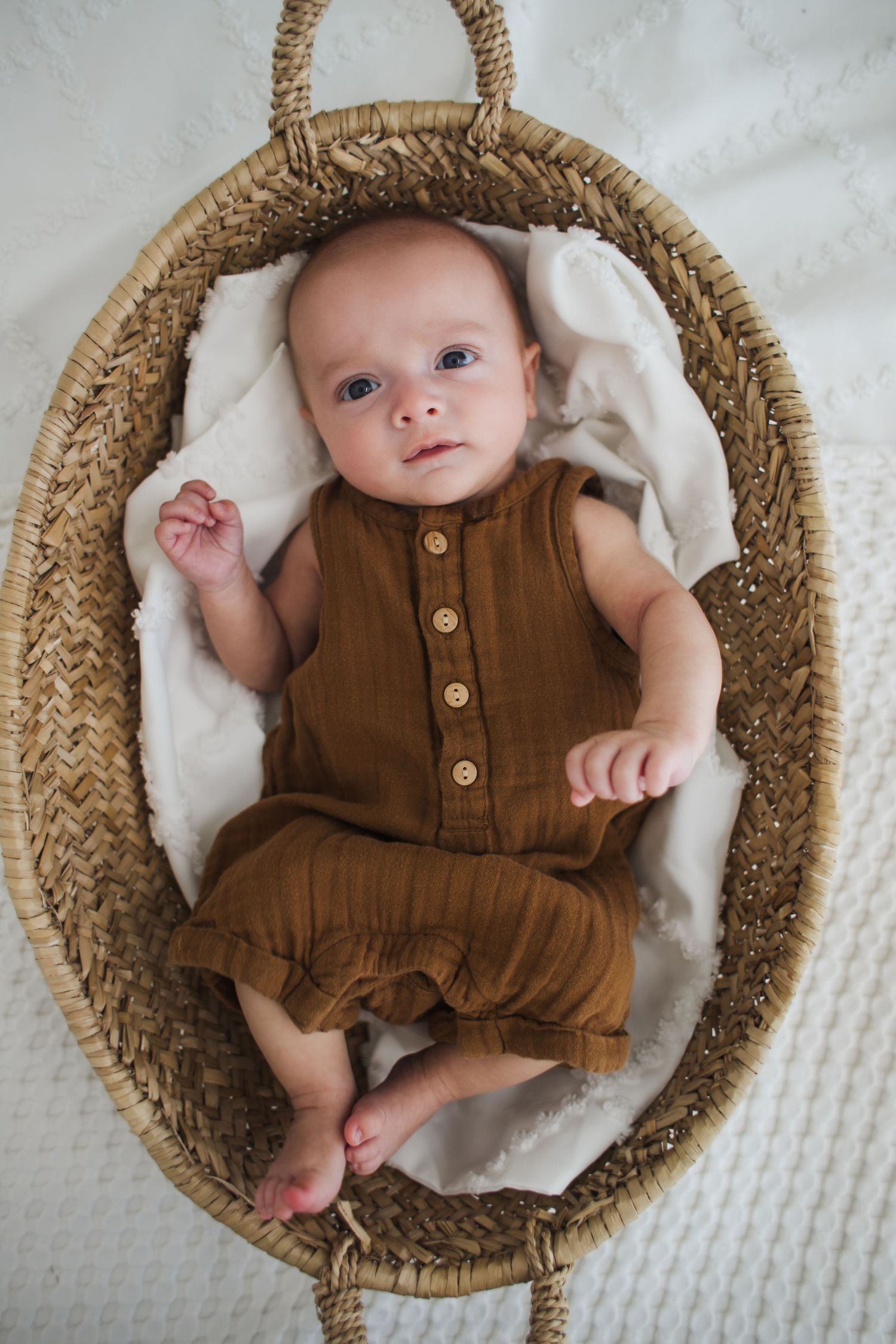 A baby wearing a brown romper lies in a round woven basket lined with white fabric, shown from different angles.